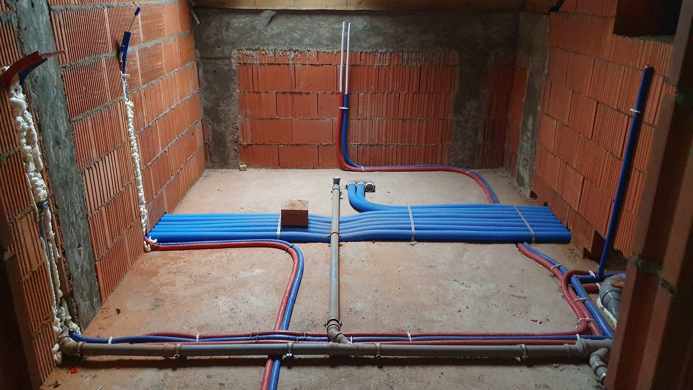 Surowy stan instalacji hydraulicznej w pomieszczeniu z ceglanymi ścianami i betonową podłogą, widoczne niebieskie rury karbowane i czerwono-niebieskie rury instalacyjne, częściowo zamontowane...