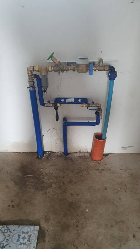 Kompletna instalacja wodna z licznikiem, zaworami i filtrem, rury w kolorze niebieskim, zamontowana przy ścianie z widocznymi śladami wilgoci na podłodze.