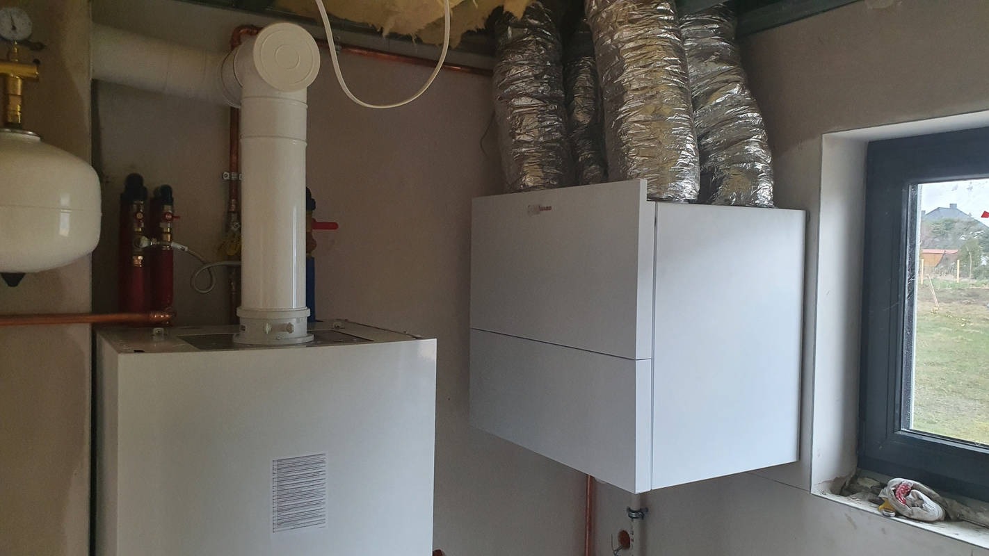 Dwa białe piece grzewcze w nowym pomieszczeniu, widoczne rury miedziane, przewody wentylacyjne i okno z widokiem na zewnątrz.