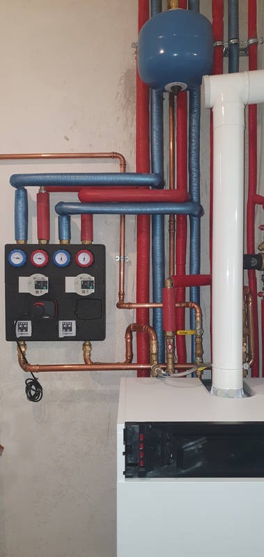 Instalacja grzewcza z kotłem, rurami miedzianymi i izolowanymi na czerwono i niebiesko, manometry na czarnej płycie, biały komin i zbiornik wyrównawczy.