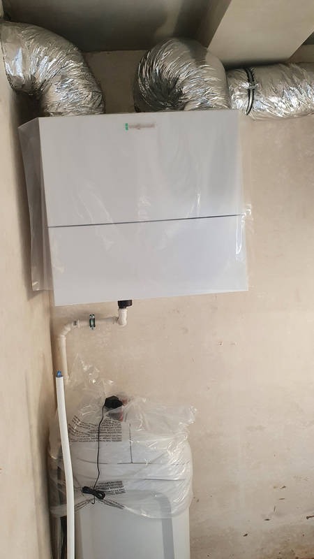 Biały piec gazowy z podłączonymi przewodami wentylacyjnymi, jeszcze w folii ochronnej, zamontowany na ścianie w surowym pomieszczeniu.