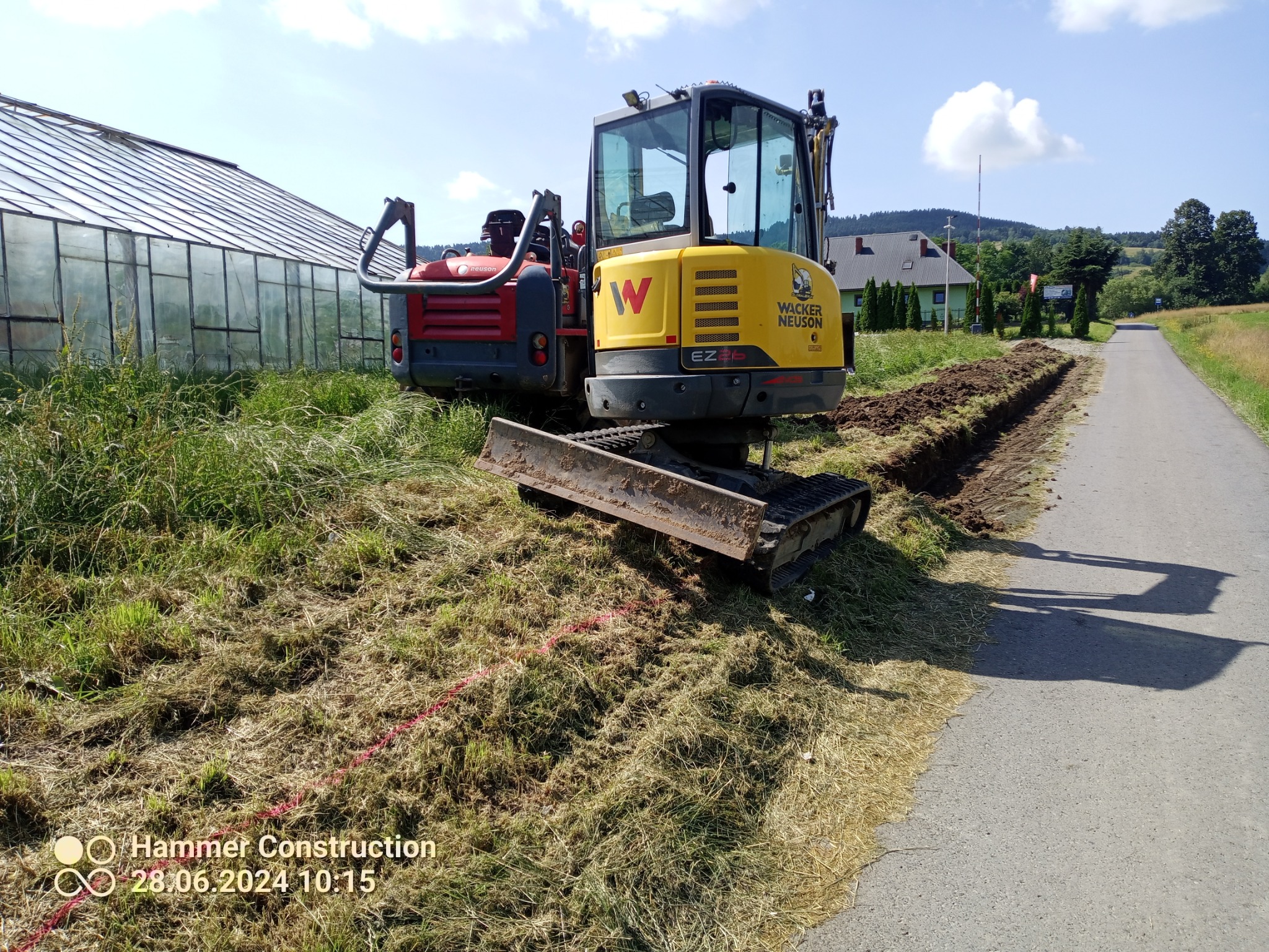 Żółto-czerwona mini koparka Wacker Neuson EZ16 na poboczu drogi, obok świeżo wykopanego rowu, z widocznym szklarnią i pagórkowatym krajobrazem w tle.