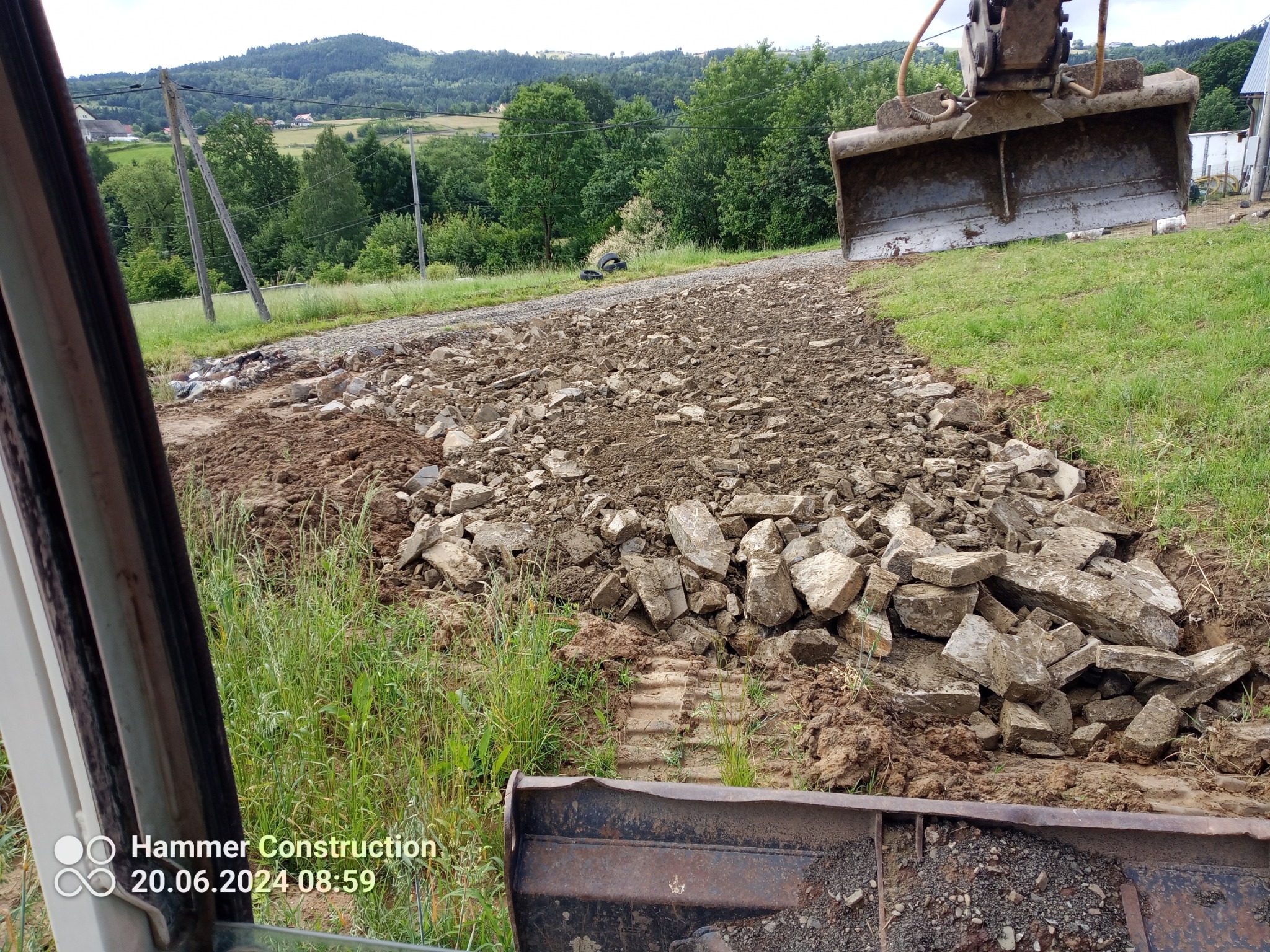 Utwardzanie drogi gruzem z użyciem koparki na tle zielonych wzgórz, widok z kabiny operatora.