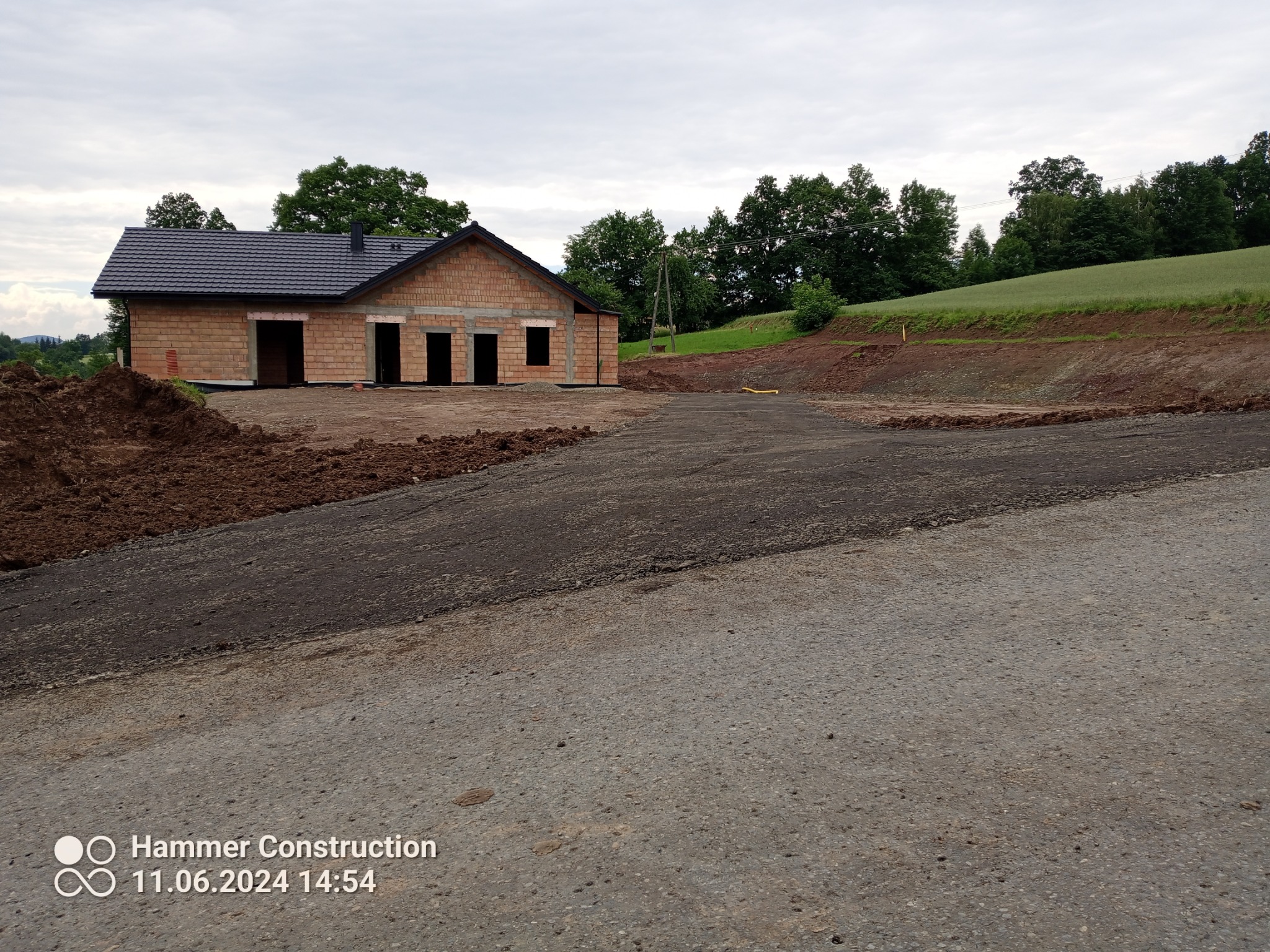 Surowy budynek jednorodzinny z ciemną dachówką, w stanie surowym otwartym, z widocznym przygotowanym i utwardzonym terenem pod drogę dojazdową, na tle zielonego wzgórza i pochmurnego nieba.