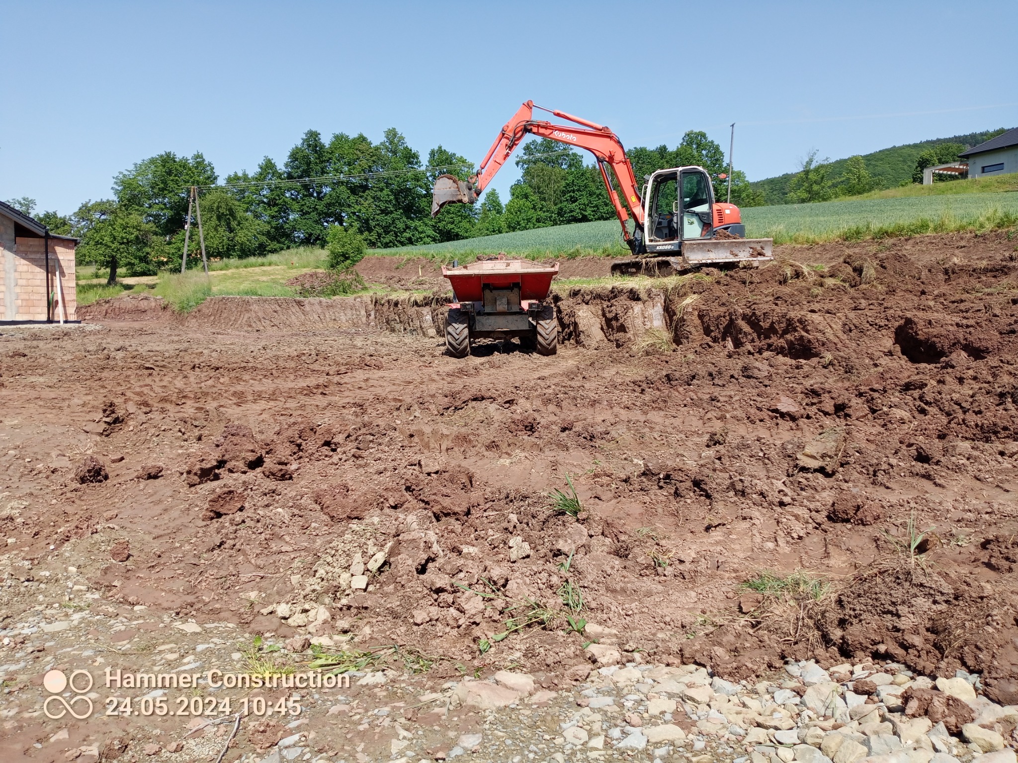 Pomarańczowa koparka Kubota nabiera ziemię do czerwonej wywrotki na placu budowy, widoczne świeże wykopy i budynek w trakcie budowy.
