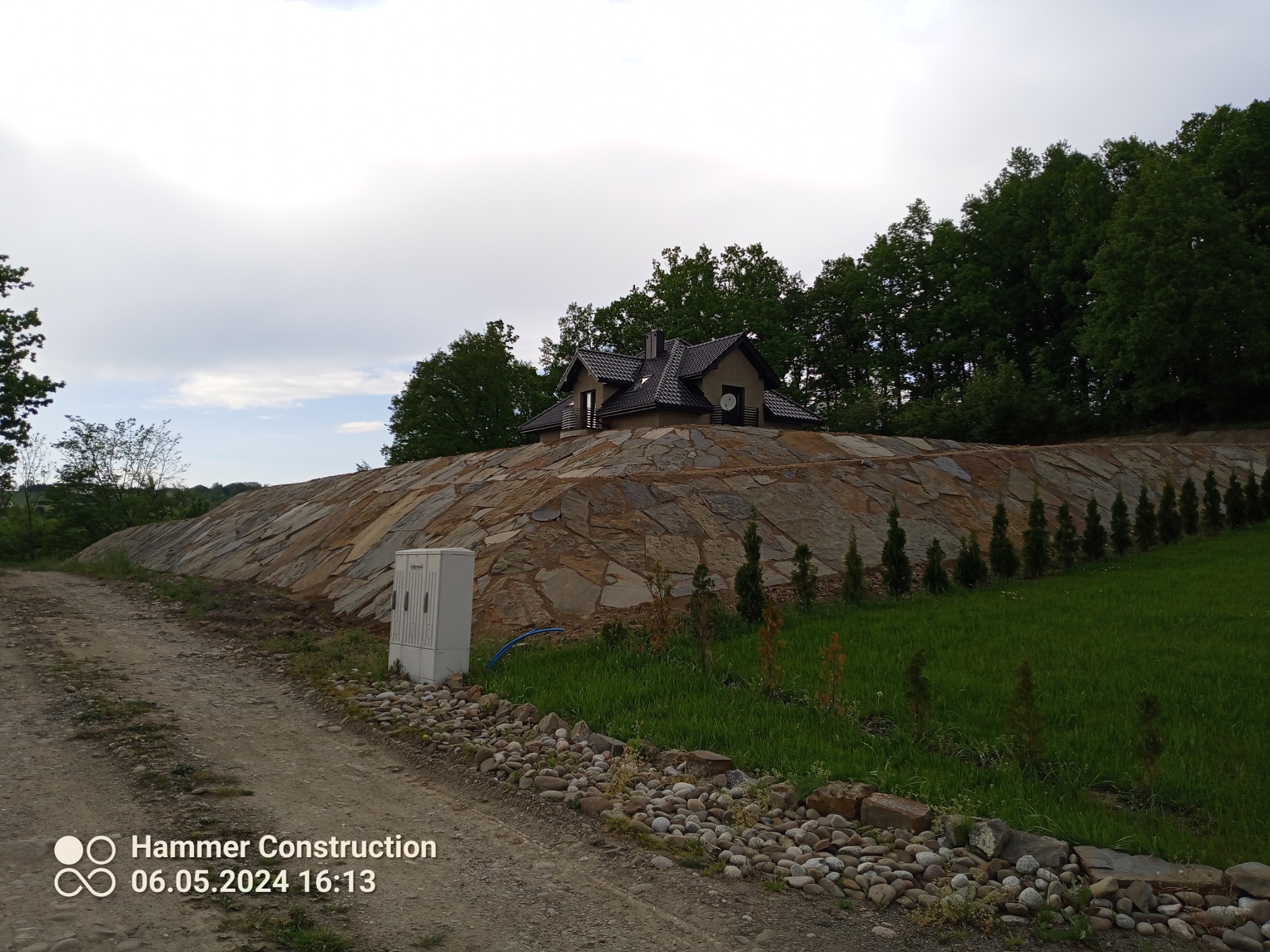 Skarpa wzmocniona kamieniami z domem na szczycie, widok z polnej drogi, małe drzewka iglaste posadzone wzdłuż zbocza.