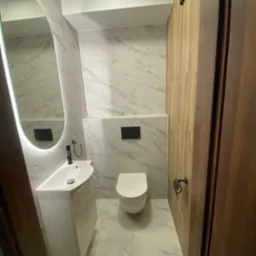 Nowoczesna łazienka z marmurowymi płytkami, podświetlanym lustrem owalnym, białą umywalką nablatową i toaletą w minimalistycznym stylu.