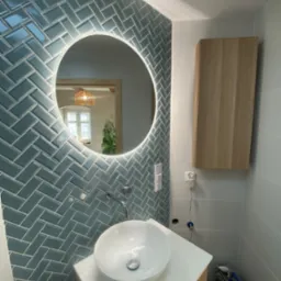 Nowoczesna łazienka z niebieską glazurą ułożoną w jodełkę, okrągłe lustro z podświetleniem LED i białą umywalką nablatową. Widoczne akcesoria i narzędzia glazurnicze.