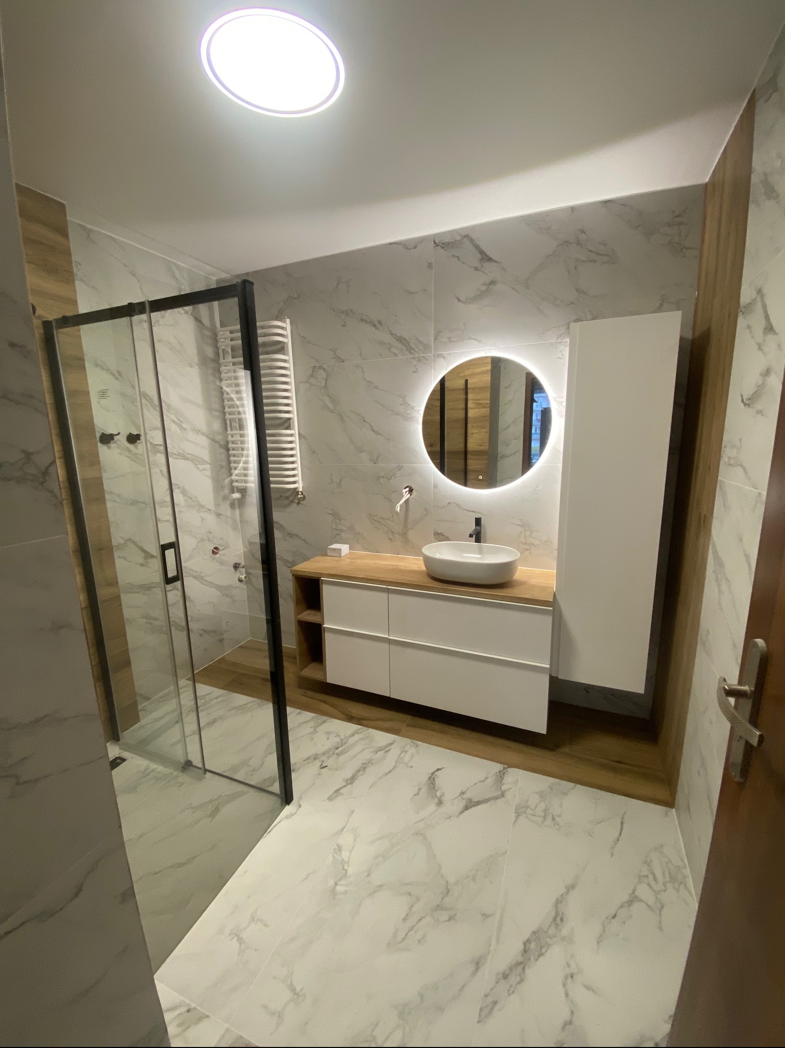 Nowoczesna łazienka z kabiną prysznicową typu walk-in, umywalką nablatową na drewnianym blacie, okrągłym lustrem z podświetleniem LED i marmurowymi płytkami.