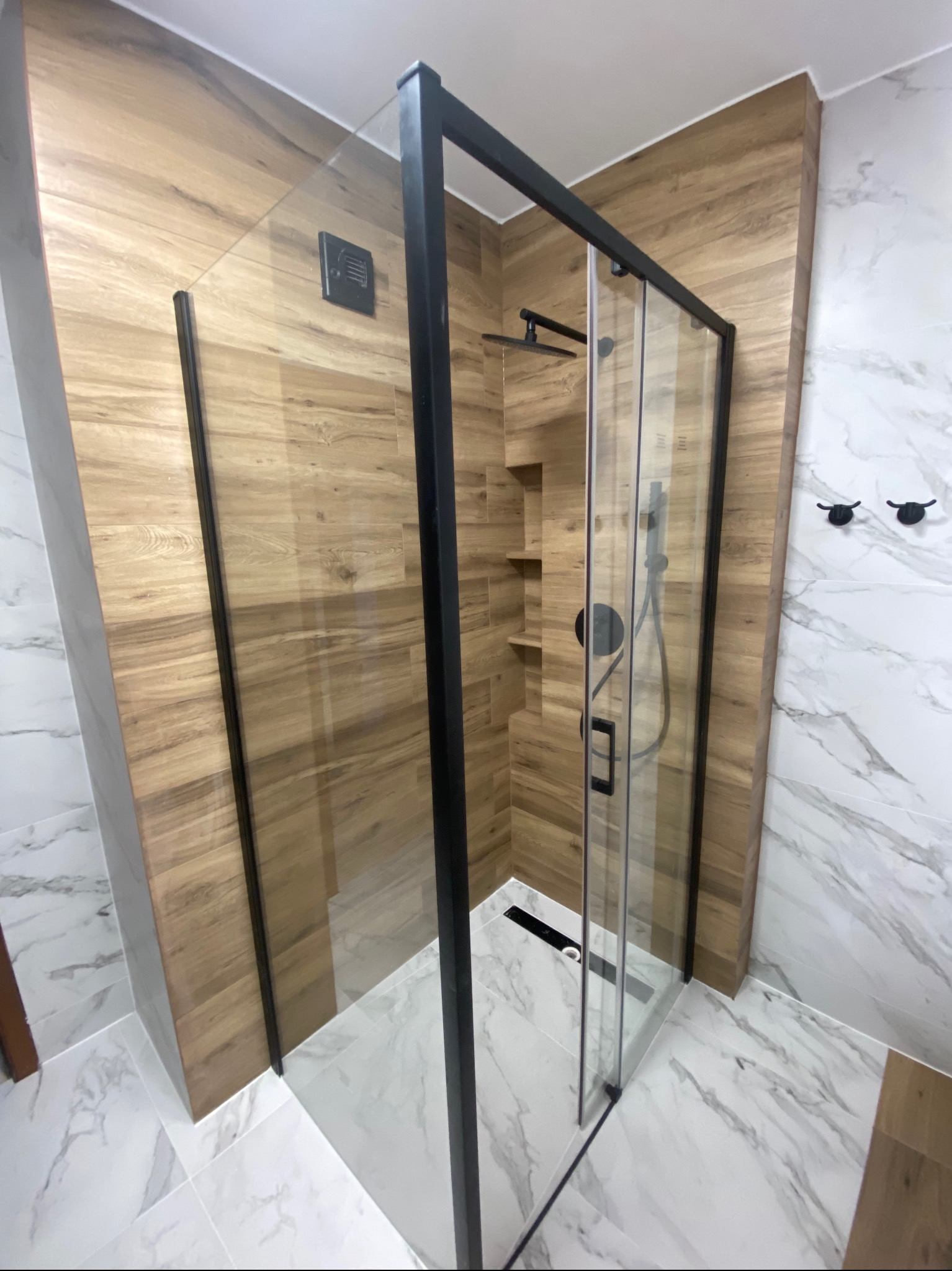 Nowoczesna kabina prysznicowa z czarnymi profilami i szklanymi drzwiami, drewnopodobne panele ścienne, marmurowe płytki podłogowe, minimalistyczne wieszaki na ręczniki.