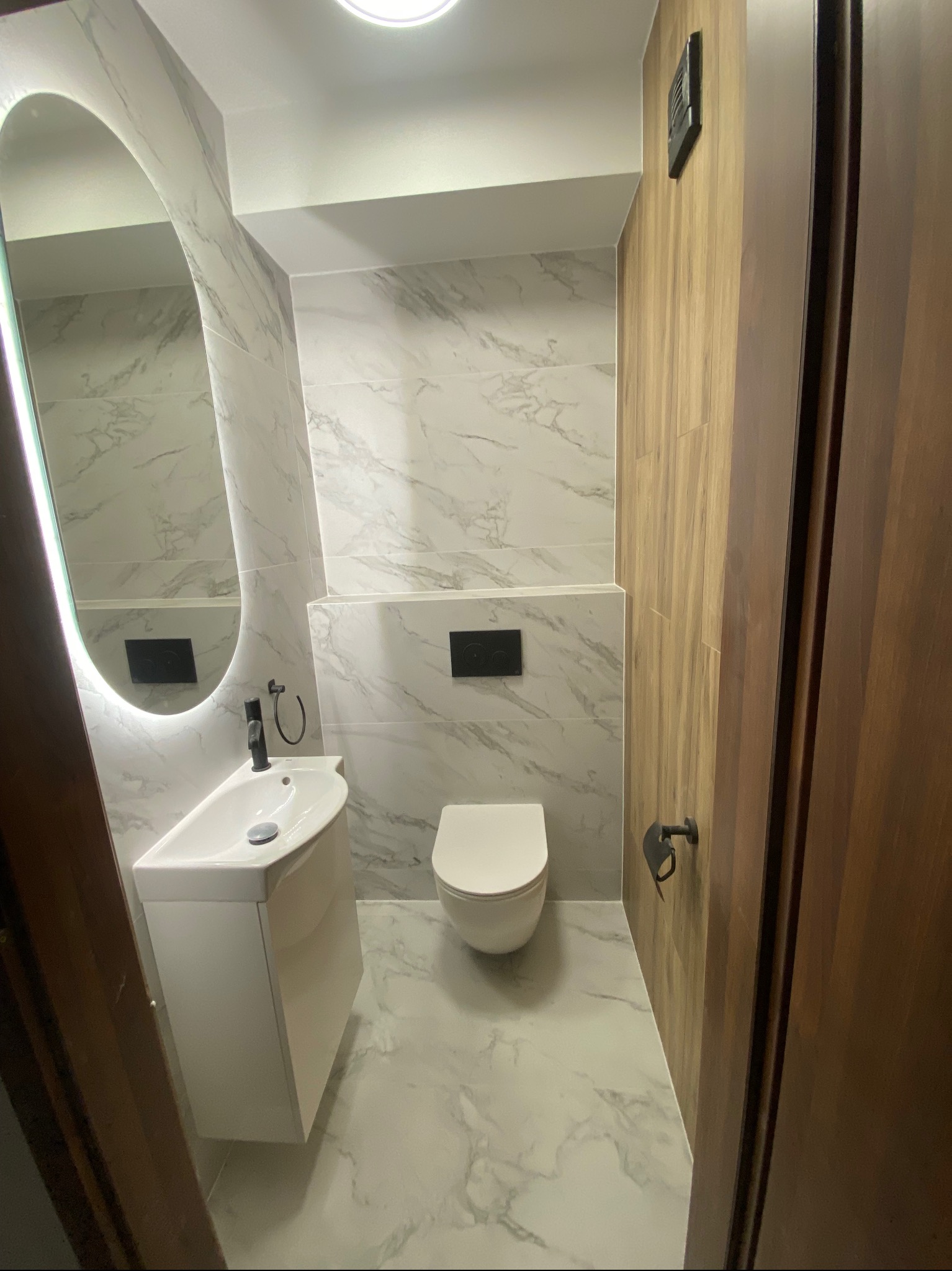 Nowoczesna łazienka z marmurowymi płytkami, podświetlanym lustrem owalnym, białą umywalką nablatową i toaletą w minimalistycznym stylu.