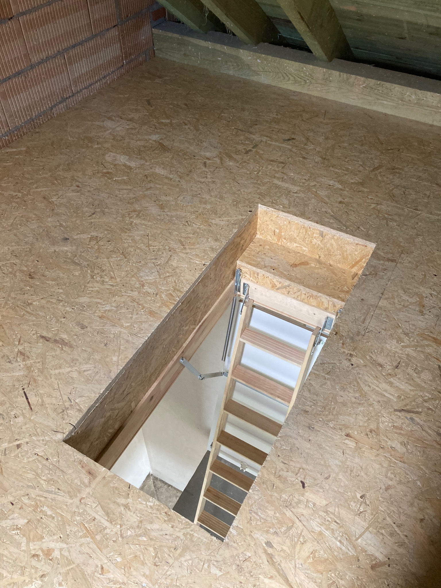 Fragment poddasza z płyt OSB z otworem i rozkładaną drabinką prowadzącą na niższy poziom, widoczne cegły i elementy konstrukcji dachu.