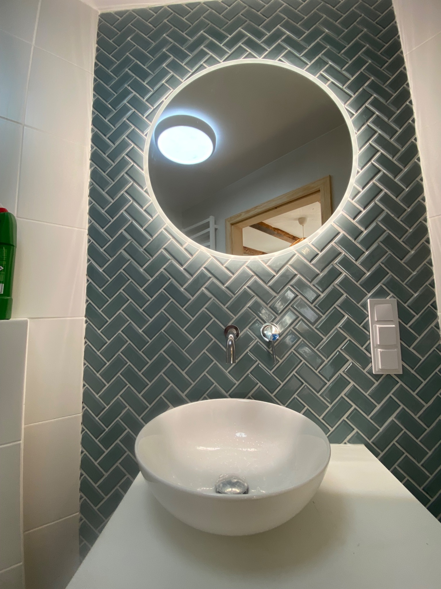 Nowoczesna łazienka z okrągłym lustrem z podświetleniem LED, umywalką nablatową i ścianą wyłożoną zielono-szarymi płytkami w jodełkę, widoczne baterie ścienne i włącznik światła.