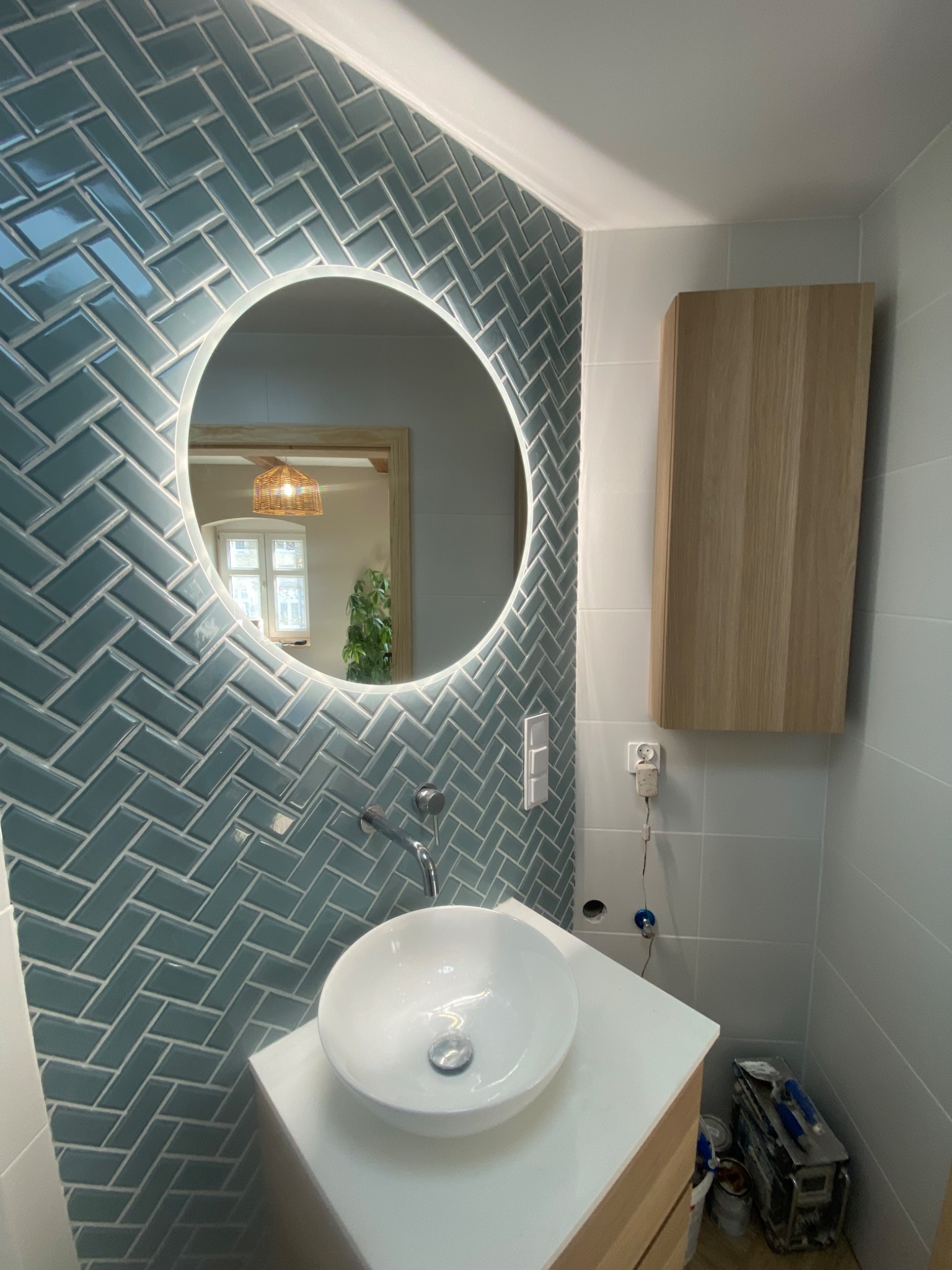 Nowoczesna łazienka z niebieską glazurą ułożoną w jodełkę, okrągłe lustro z podświetleniem LED i białą umywalką nablatową. Widoczne akcesoria i narzędzia glazurnicze.