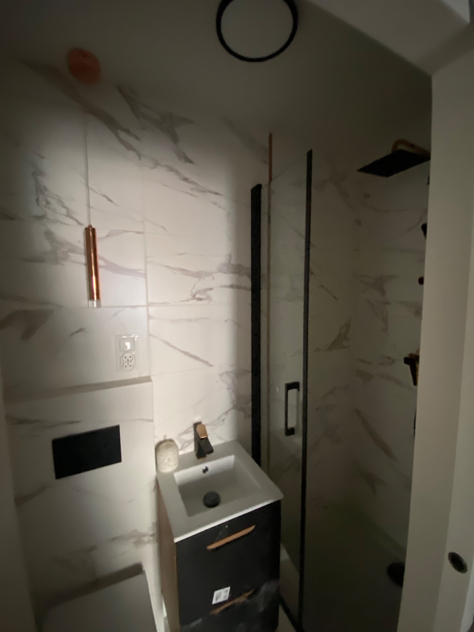 Nowoczesna łazienka z marmurowymi płytkami, czarną armaturą i kabiną prysznicową typu walk-in z czarnymi profilami, oświetlona wiszącą lampą w kolorze miedzi.