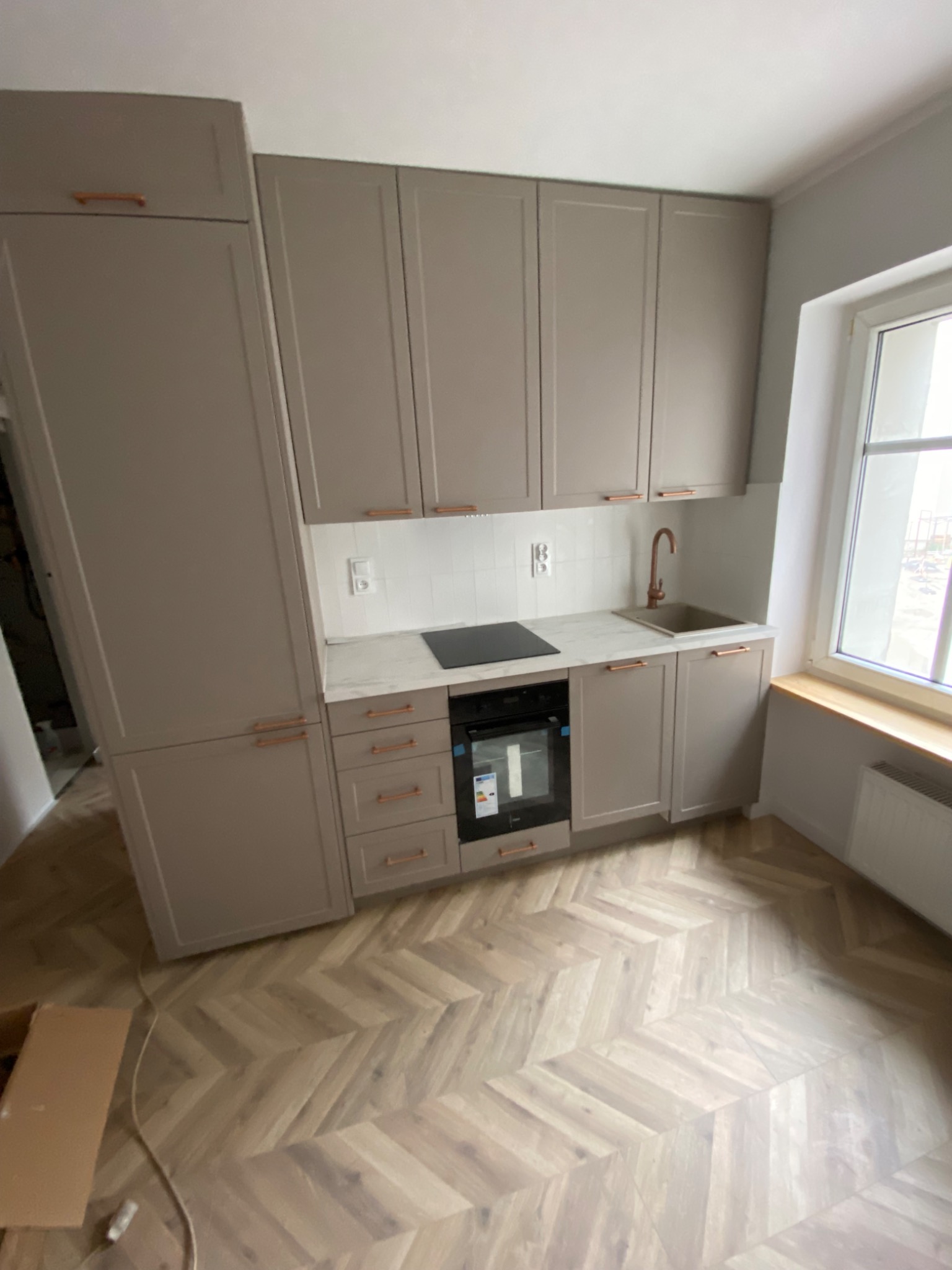 Nowoczesna kuchnia w odcieniach szarości z drewnianą podłogą w jodełkę, miedziane uchwyty szafek, widoczny fragment okna i grzejnika.