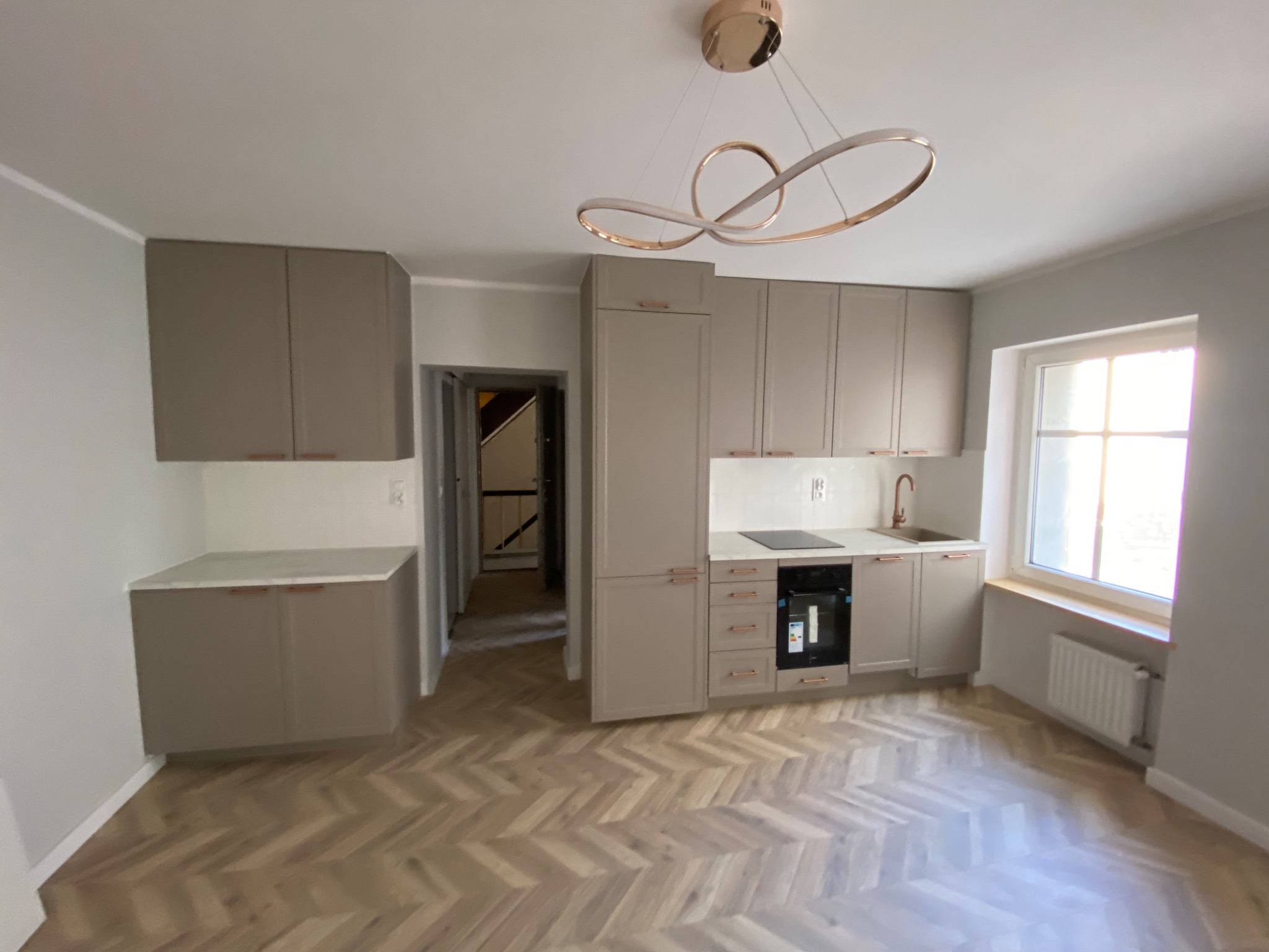 Jasna kuchnia z szarymi szafkami, parkietem w jodełkę i nowoczesną lampą sufitową w kształcie abstrakcyjnych okręgów.
