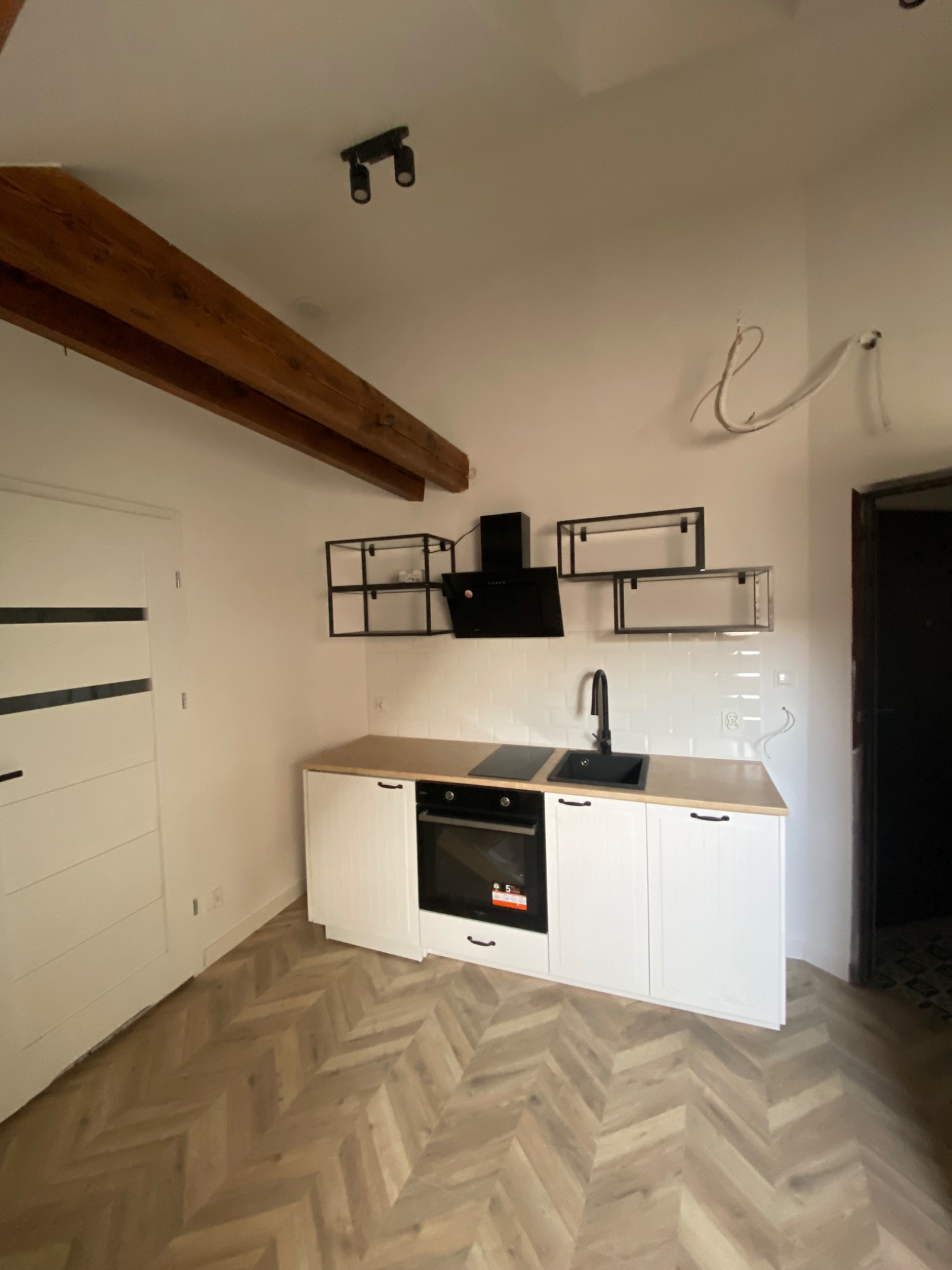 Nowoczesna kuchnia z białymi szafkami, drewnianym blatem i czarnymi akcentami, w tym okapem i baterią, uzupełniona o drewnianą belkę stropową i parkiet ułożony w jodełkę.