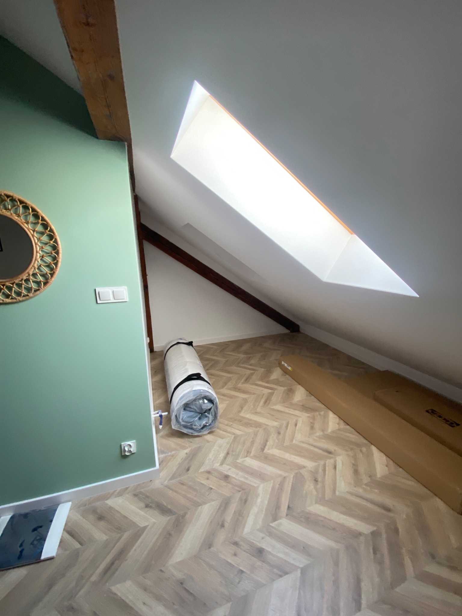 Poddasze po remoncie: ściana pomalowana na zielono, okrągłe lustro na ścianie, parkiet ułożony w jodełkę, zwinięty materac w folii i kartonowe opakowanie, okno dachowe w skosie.