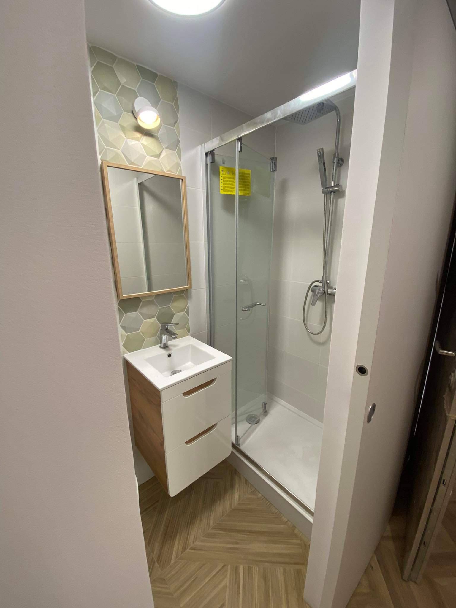 Nowoczesna łazienka po remoncie: kabina prysznicowa z deszczownicą, umywalka z szafką w kolorze drewna, lustro z podświetleniem i heksagonalne płytki na ścianie.