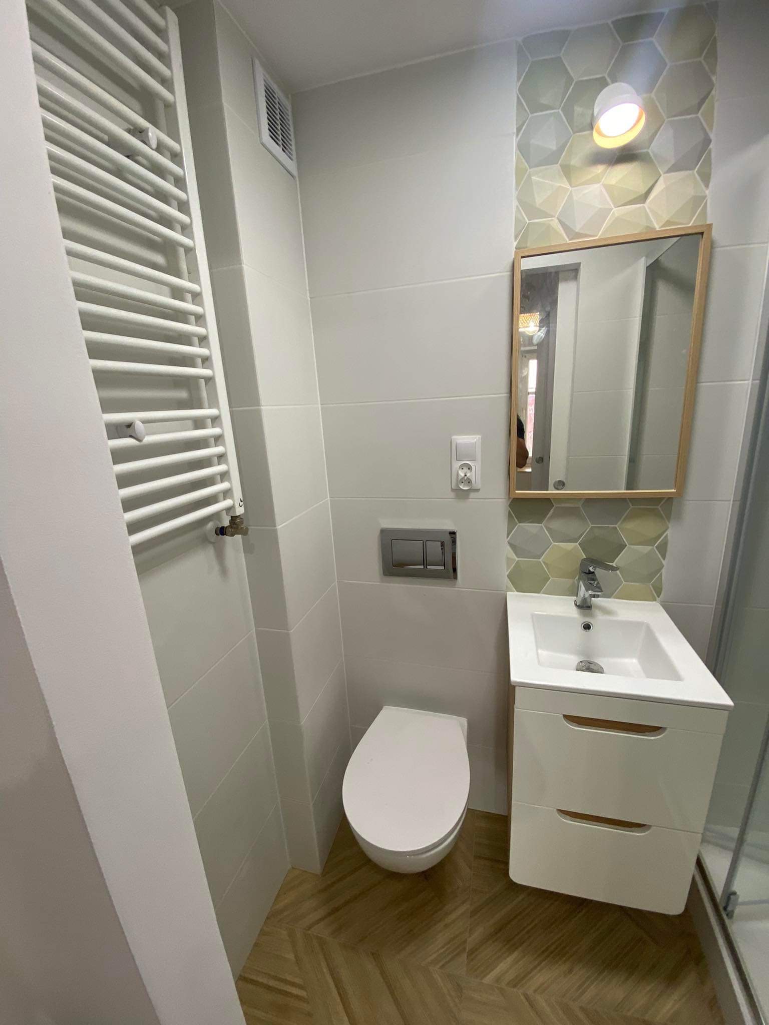 Nowoczesna łazienka z białą armaturą, grzejnikiem drabinkowym, szafką pod umywalką z drewnianymi akcentami i heksagonalnymi płytkami dekoracyjnymi nad lustrem.