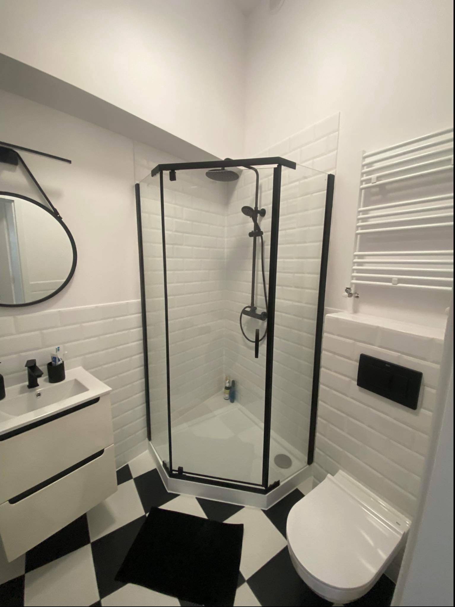 Nowoczesna łazienka w Czempiniu z kabiną prysznicową w czarnej ramie, białą armaturą i czarno-białą szachownicą na podłodze, uzupełniona okrągłym lustrem i grzejnikiem drabinkowym.