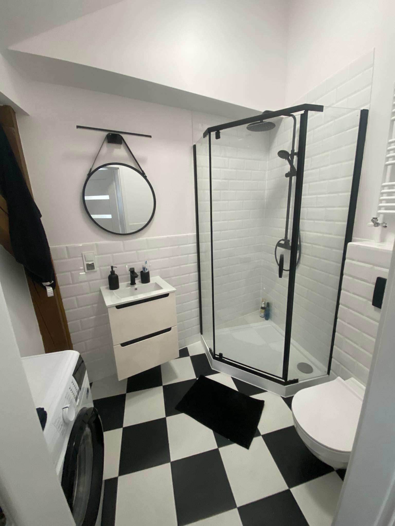 Nowoczesna łazienka z czarno-białą szachownicą na podłodze, kabiną prysznicową z czarną ramą, okrągłym lustrem na ścianie z białych płytek imitujących cegłę oraz białą szafką pod umywalką.