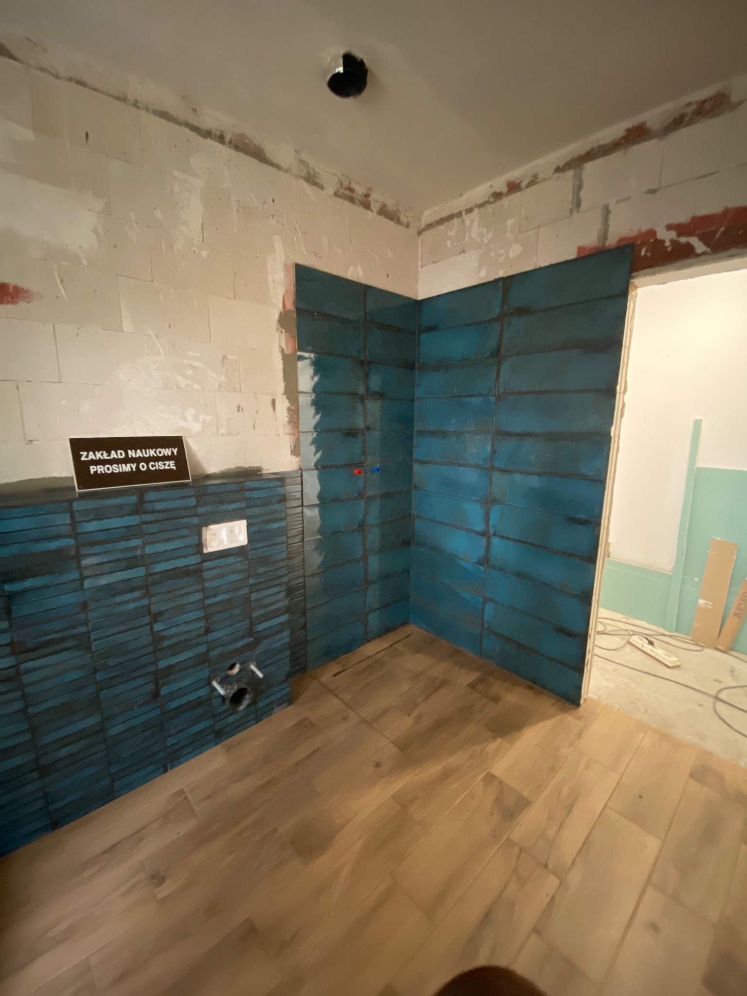 Niebiesko-czarne płytki ułożone na ścianie łazienki w trakcie remontu, widoczny fragment drewnianej podłogi, niezabezpieczone kable i rury oraz tabliczka z napisem 'Zakład naukowy prosimy o ciszę'.