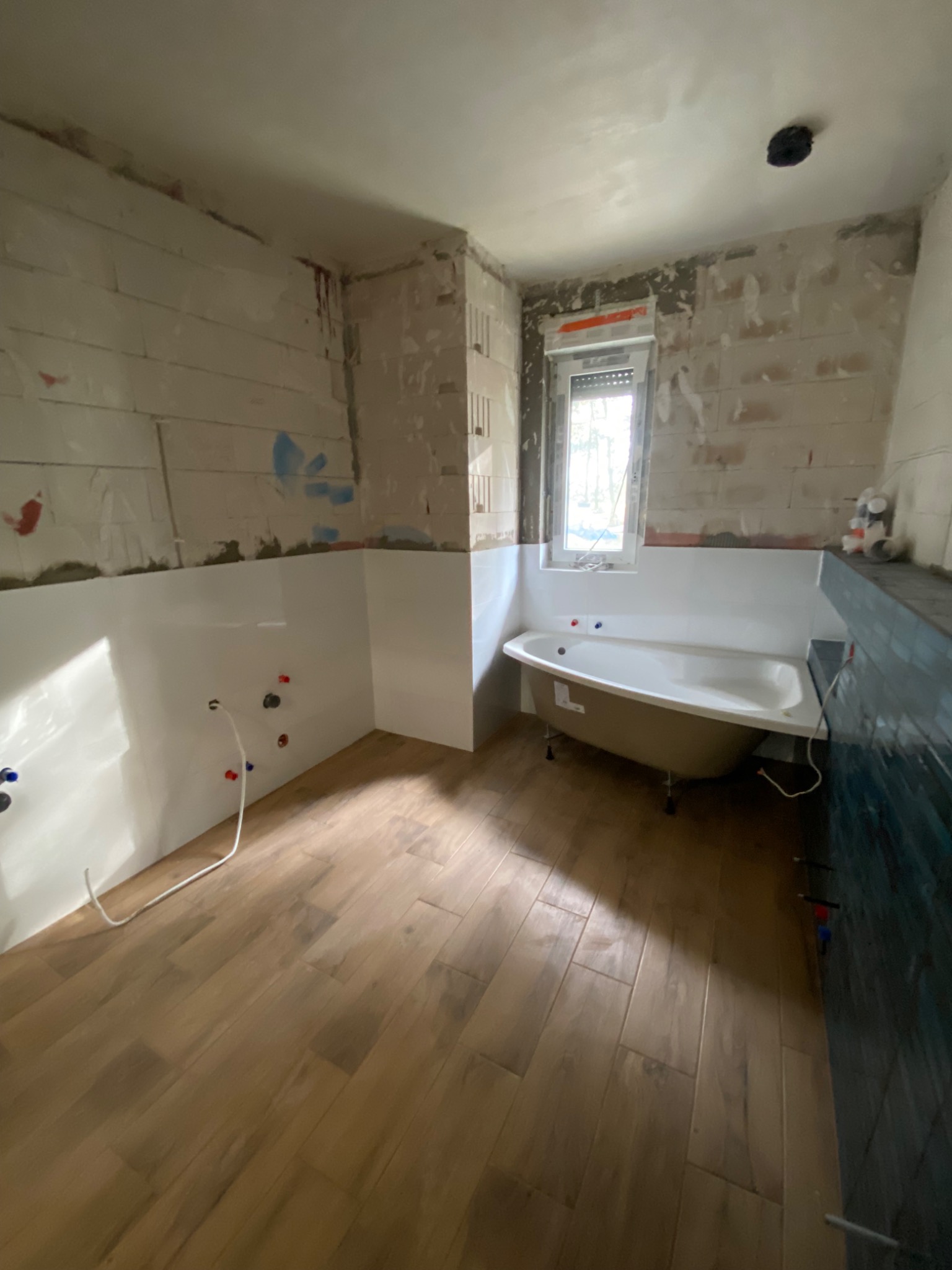 Łazienka w trakcie prac: częściowo wykończone ściany z białymi panelami, odsłonięte cegły, drewnopodobna podłoga, wanna na nóżkach, okno z roletą.