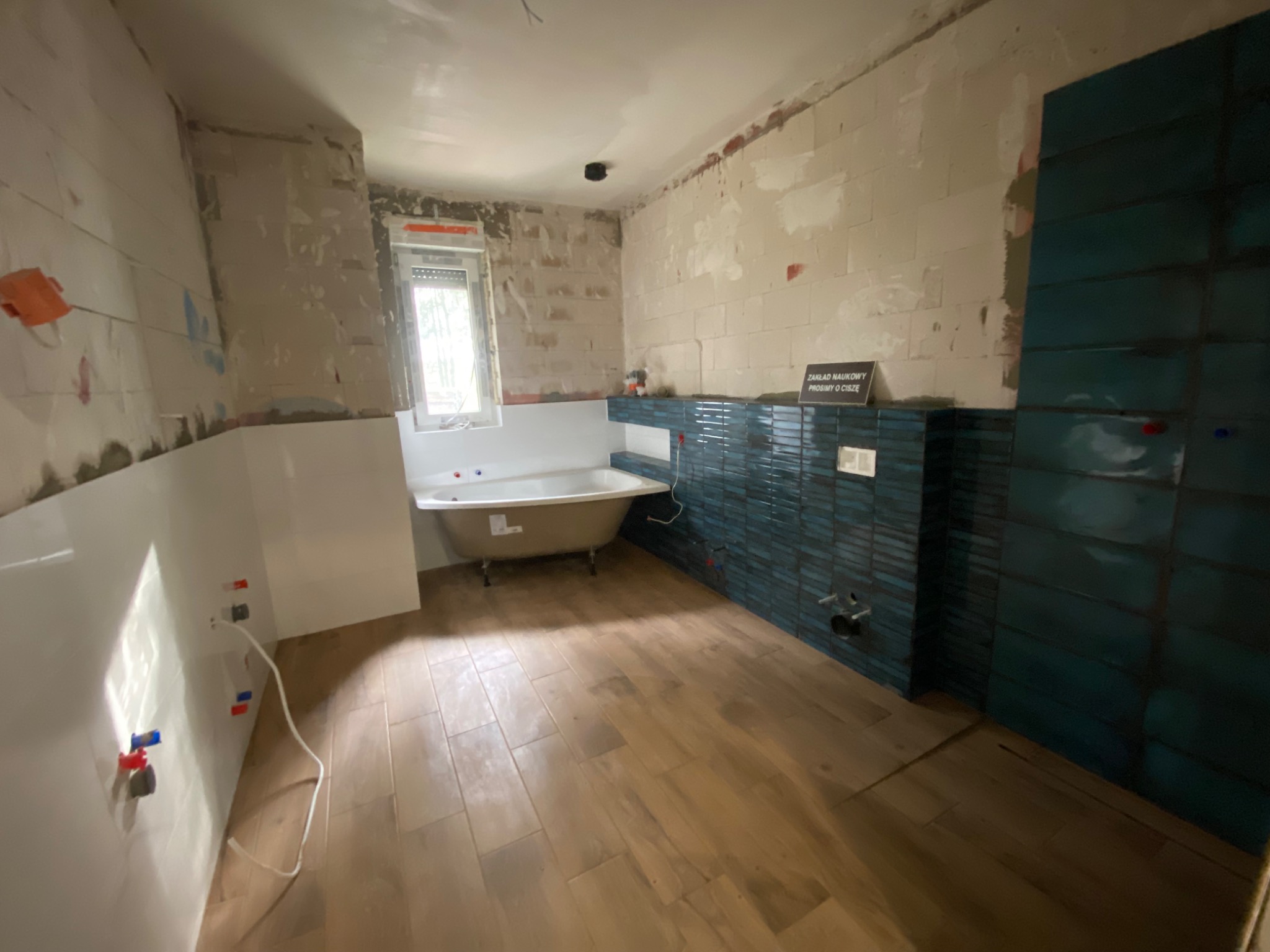 Wnętrze łazienki w trakcie remontu: częściowo położone turkusowe płytki na ścianie, drewnopodobna podłoga, wanna na nóżkach, ściany przygotowane do dalszych prac wykończeniowych.
