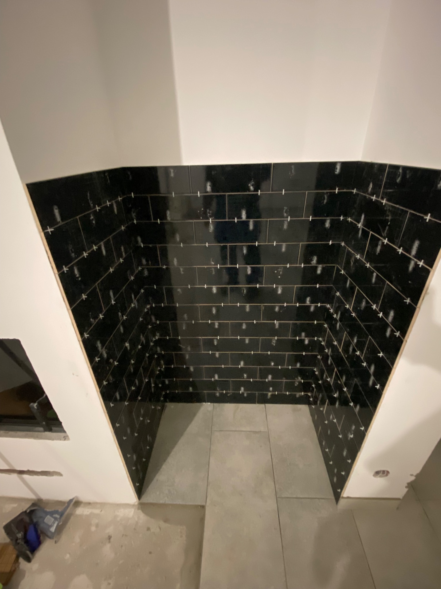 Fragment łazienki w trakcie układania czarnych płytek typu cegiełka z białymi krzyżykami dystansowymi oraz szare płytki podłogowe, widoczne narzędzia i elementy wykończeniowe.