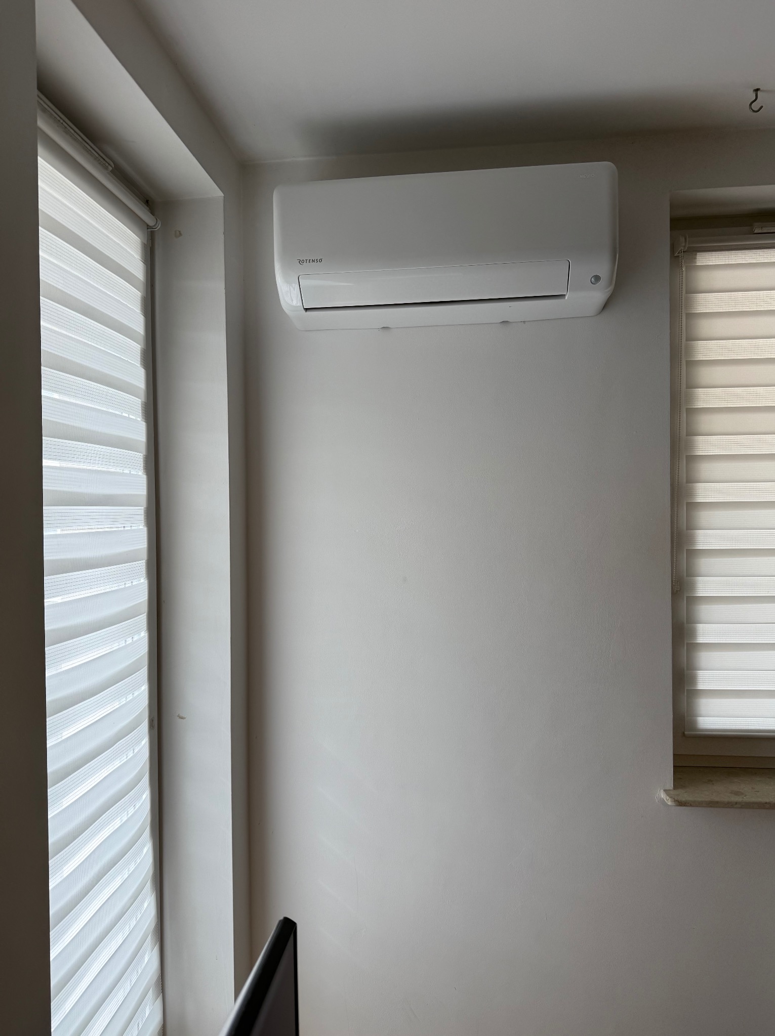 Biały klimatyzator ROTENSO zamontowany na ścianie w jasnym pomieszczeniu, widoczne rolety okienne.