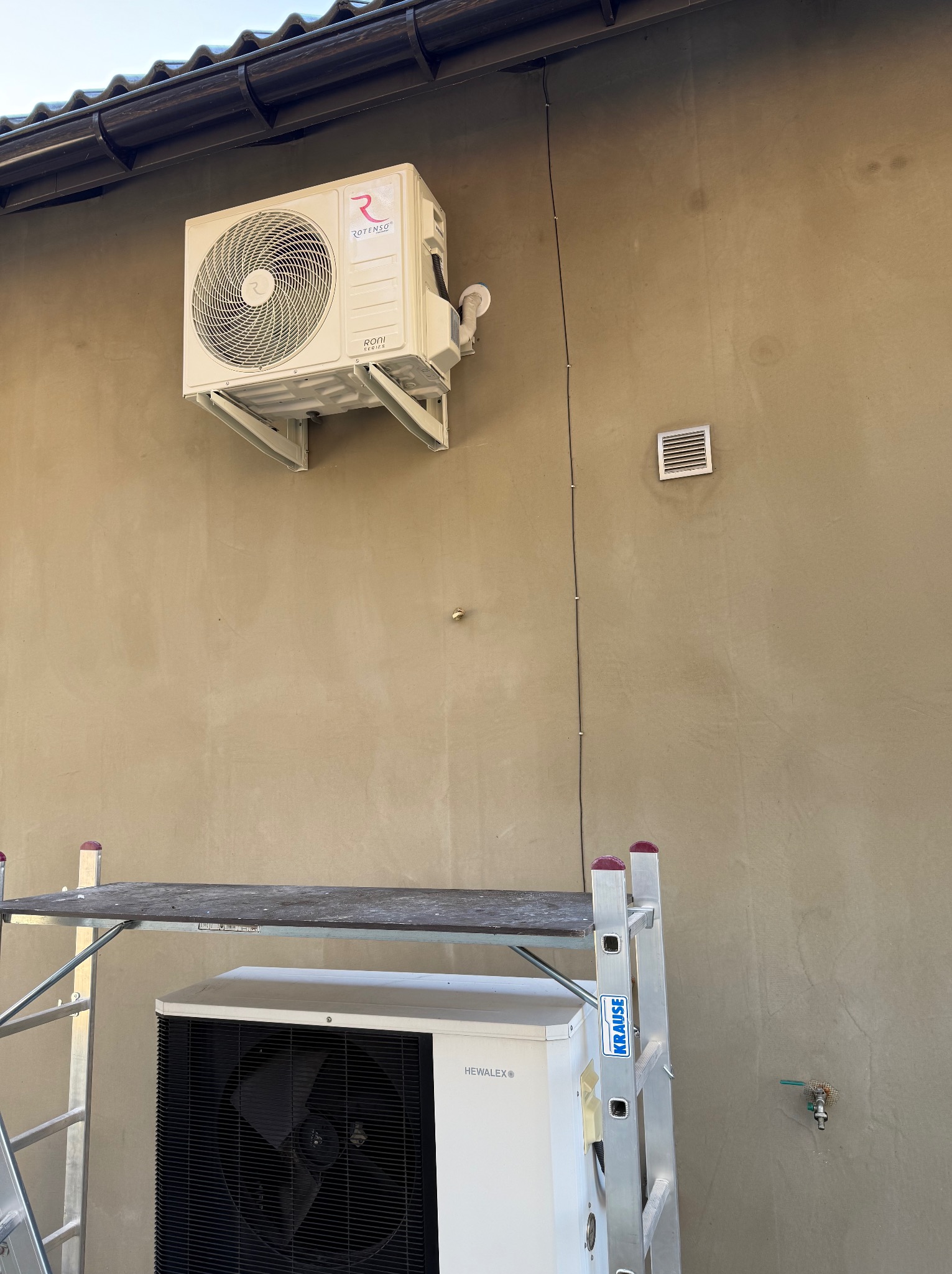 Zewnętrzna jednostka klimatyzacji Rotenso zamontowana na wspornikach, widoczna drabina Krause i dolna część jednostki zewnętrznej Hewalex, brązowa elewacja budynku.