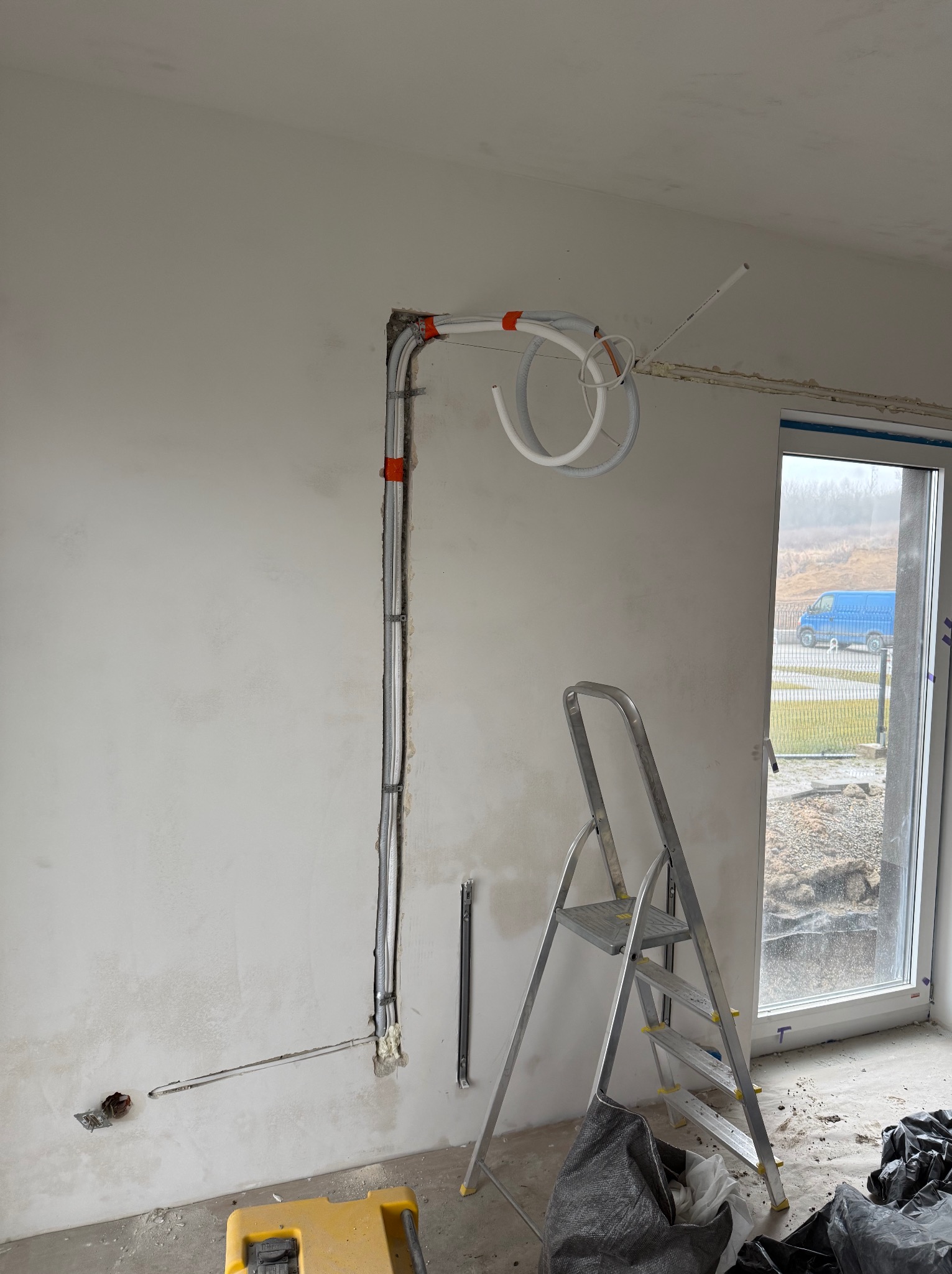 Wewnętrzna instalacja rur wentylacyjnych i przewodów elektrycznych w surowym pomieszczeniu, widoczna drabina aluminiowa i fragment okna z widokiem na zewnątrz.