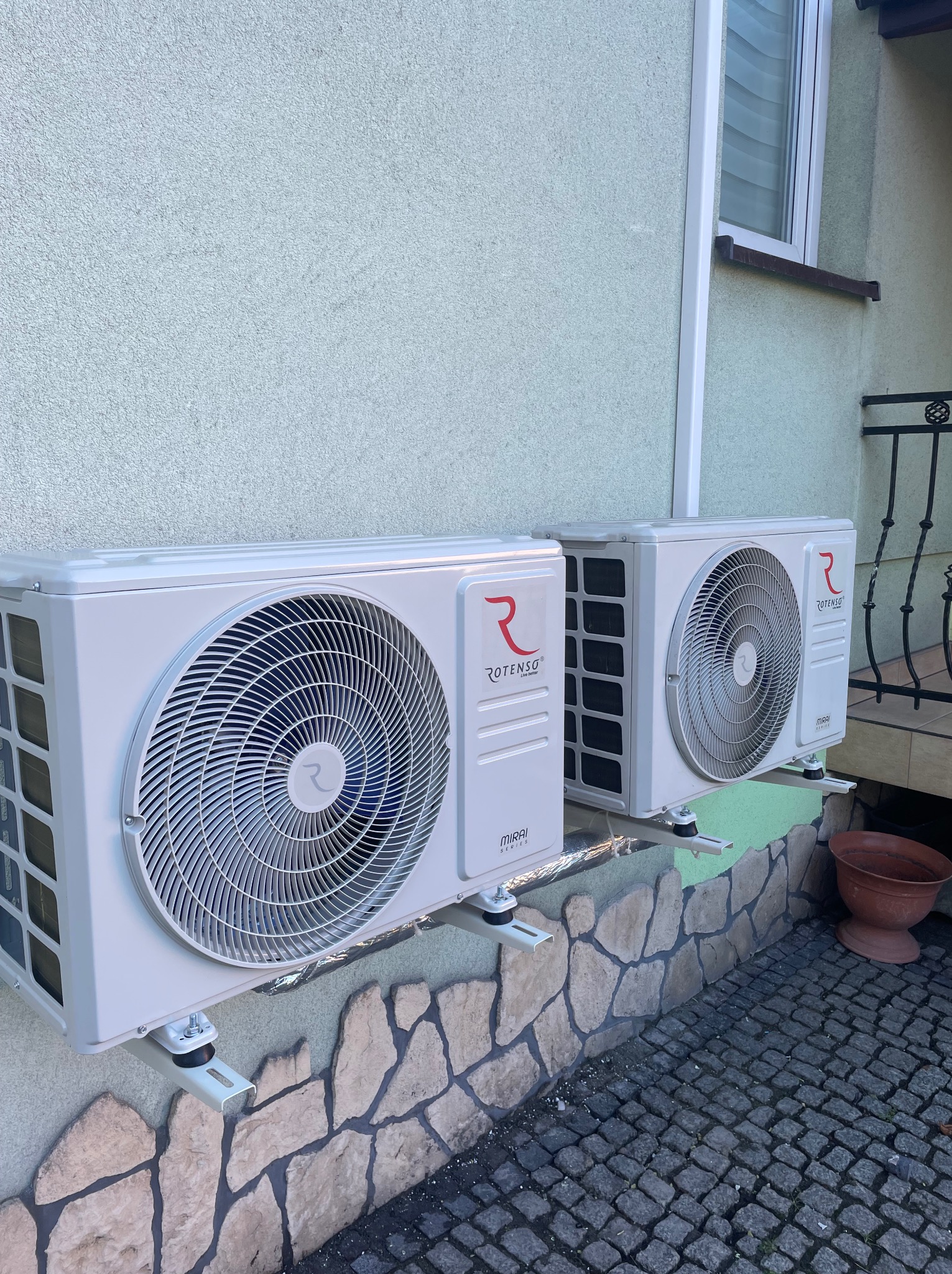 Dwa białe klimatyzatory Rotenso Mirai Series zamontowane na elewacji domu z podmurówką z kamienia, widoczne wsporniki montażowe i kostka brukowa.