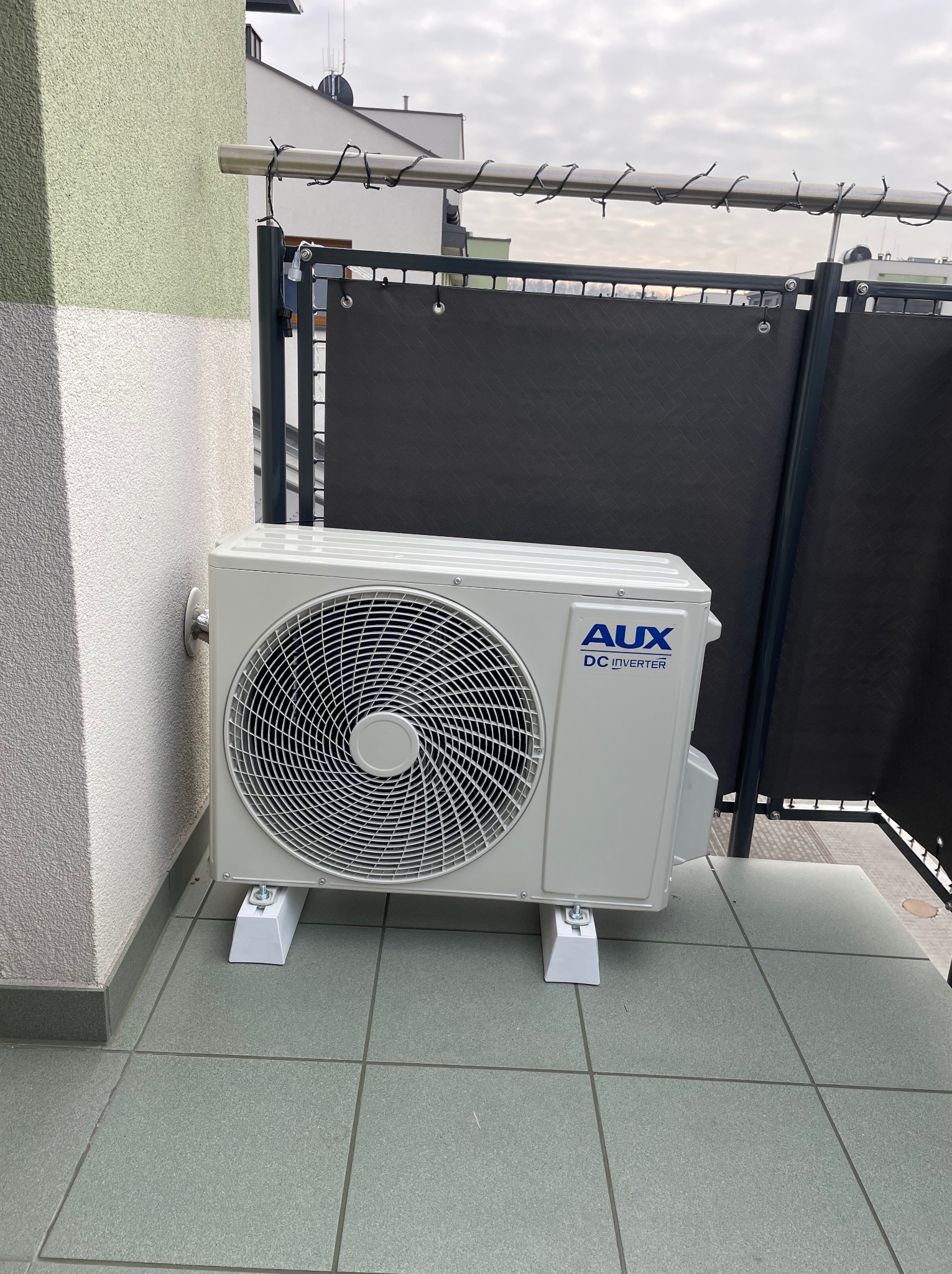 Biała jednostka zewnętrzna klimatyzatora AUX DC Inverter, zamontowana na balkonie na podwyższeniach, widok z boku, szara podłoga z płytek.