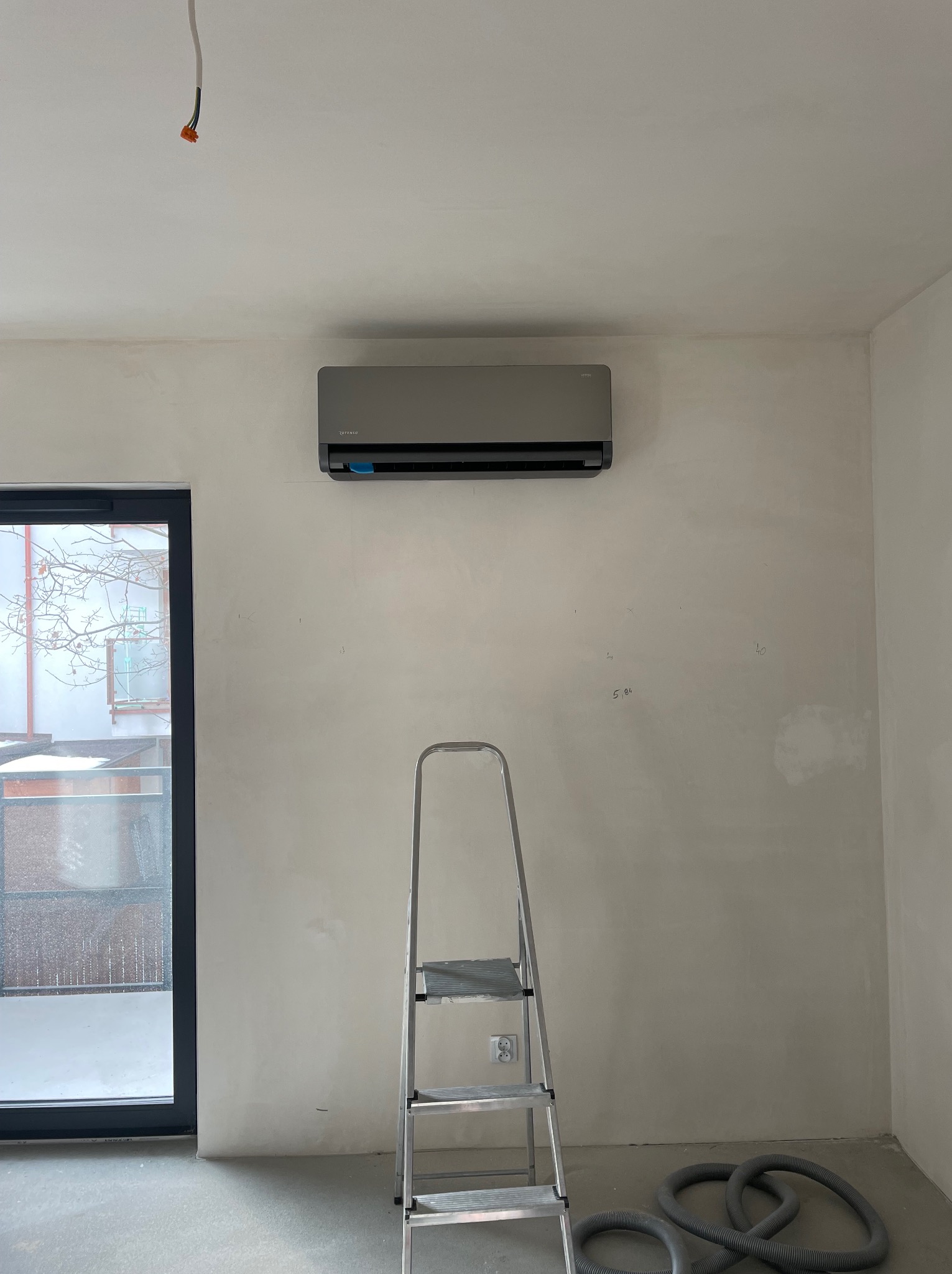 Szary klimatyzator zamontowany na ścianie w surowym wnętrzu, widoczna drabina i wąż wentylacyjny na podłodze, instalacja elektryczna wystająca z sufitu.