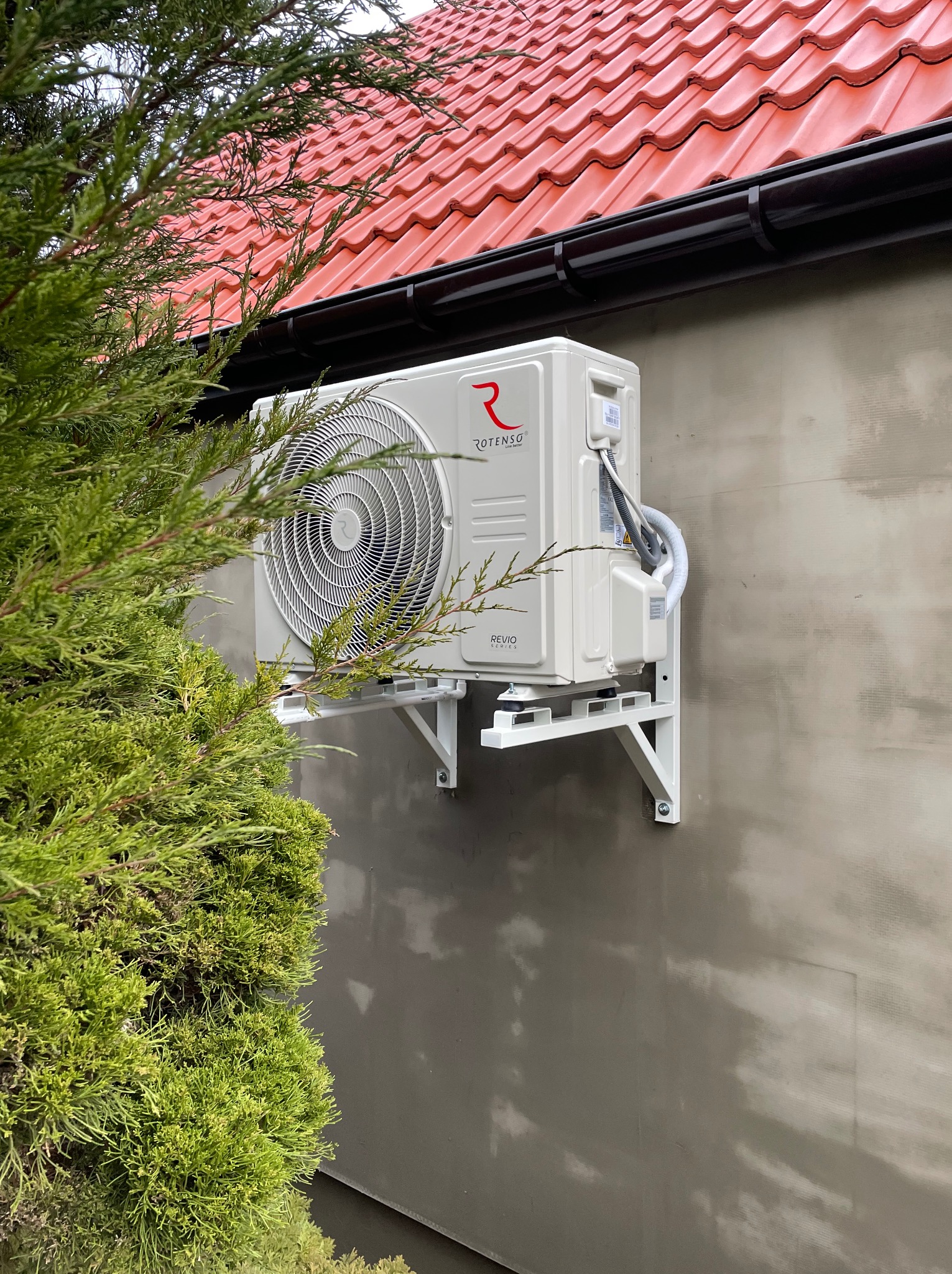 Zewnętrzna jednostka klimatyzacji Rotenso zamontowana na białym wsporniku na szarej ścianie budynku, z widocznymi kablami i rurami, częściowo zasłonięta przez zielone gałęzie drzewa iglastego...
