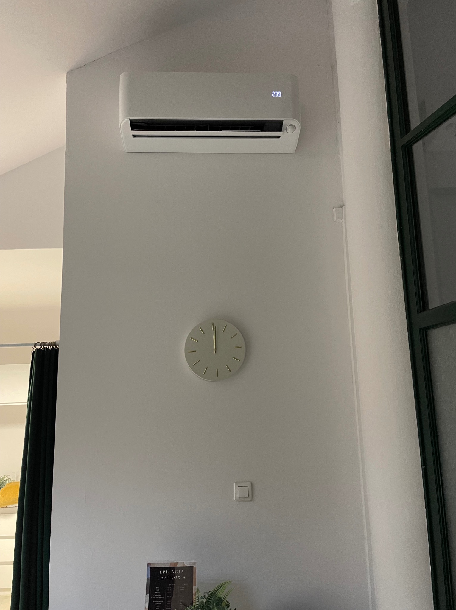 Biały klimatyzator ścienny z wyświetlaczem temperatury 23.9 stopni Celsjusza, zamontowany nad okrągłym zegarem na białej ścianie w jasnym pomieszczeniu.