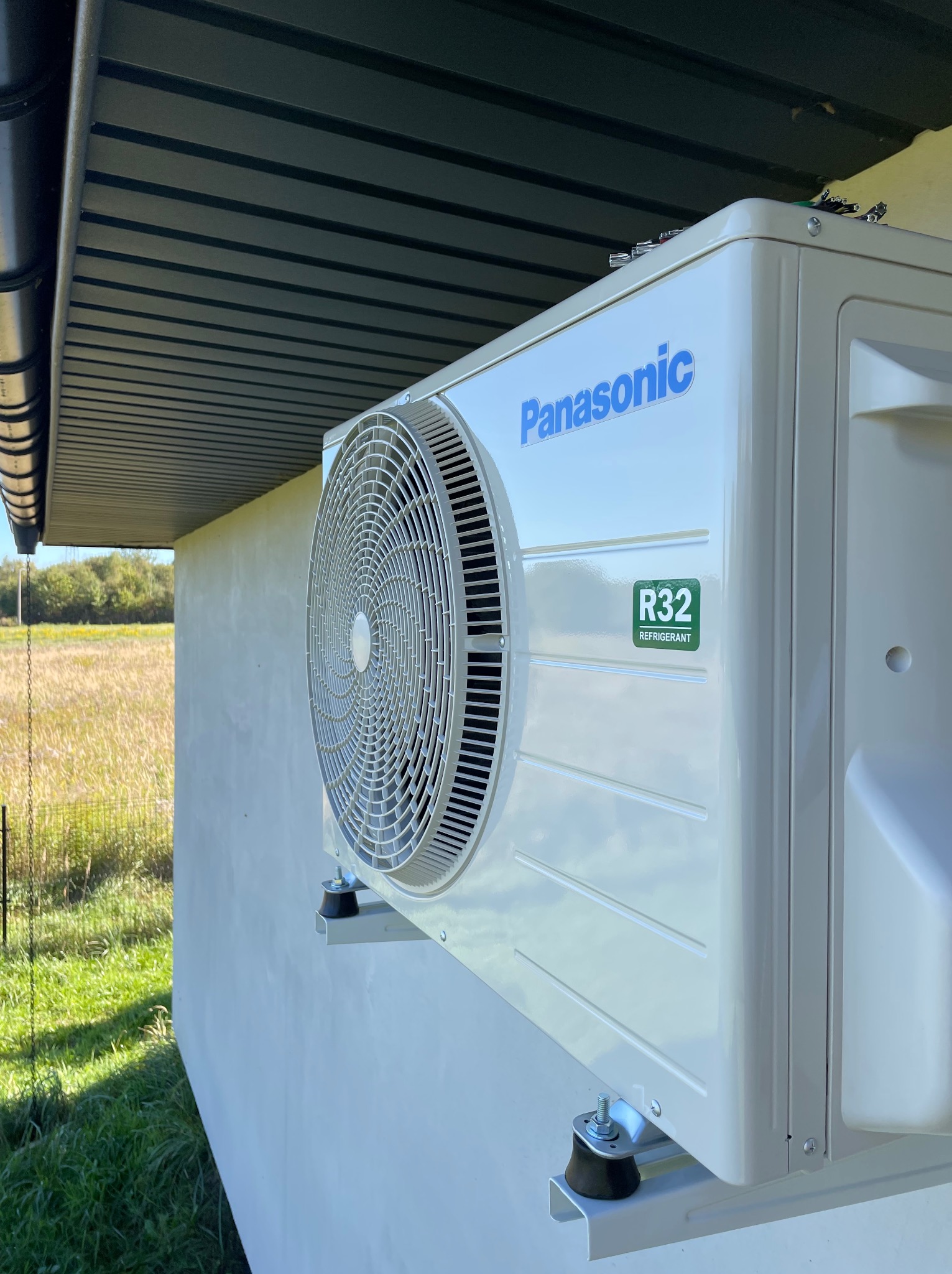 Zewnętrzna jednostka klimatyzacji Panasonic zamontowana na ścianie budynku, widok z boku, z widocznym logo i oznaczeniem czynnika chłodniczego R32, na tle pola i zieleni.