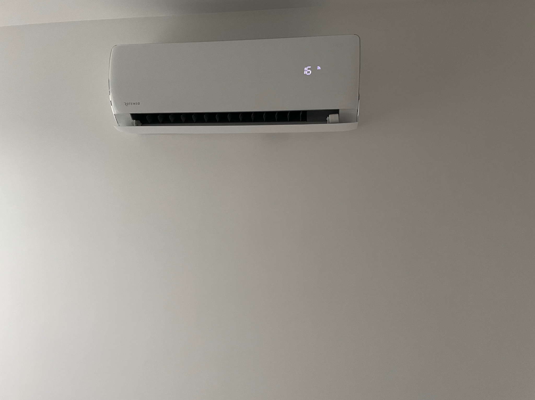 Biały klimatyzator marki Rotenso zamontowany na szarej ścianie, wyświetlający temperaturę 16 stopni.