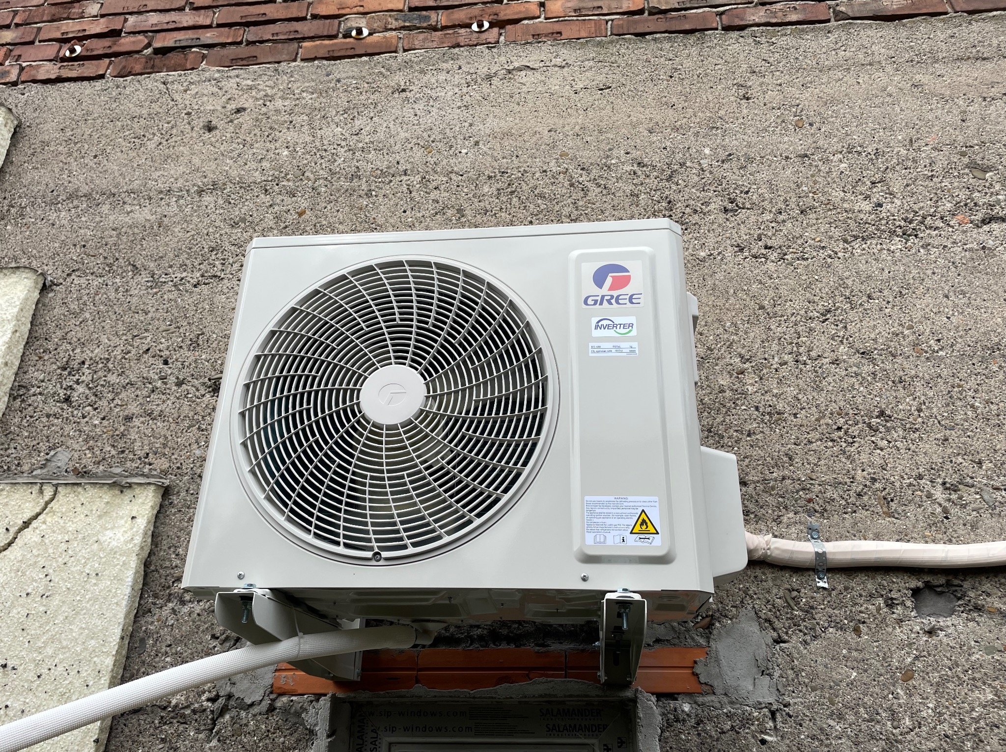 Zewnętrzna jednostka klimatyzacji marki Gree zamontowana na szarej, betonowej ścianie budynku z widocznymi elementami instalacji i fragmentem ceglanego muru u góry.