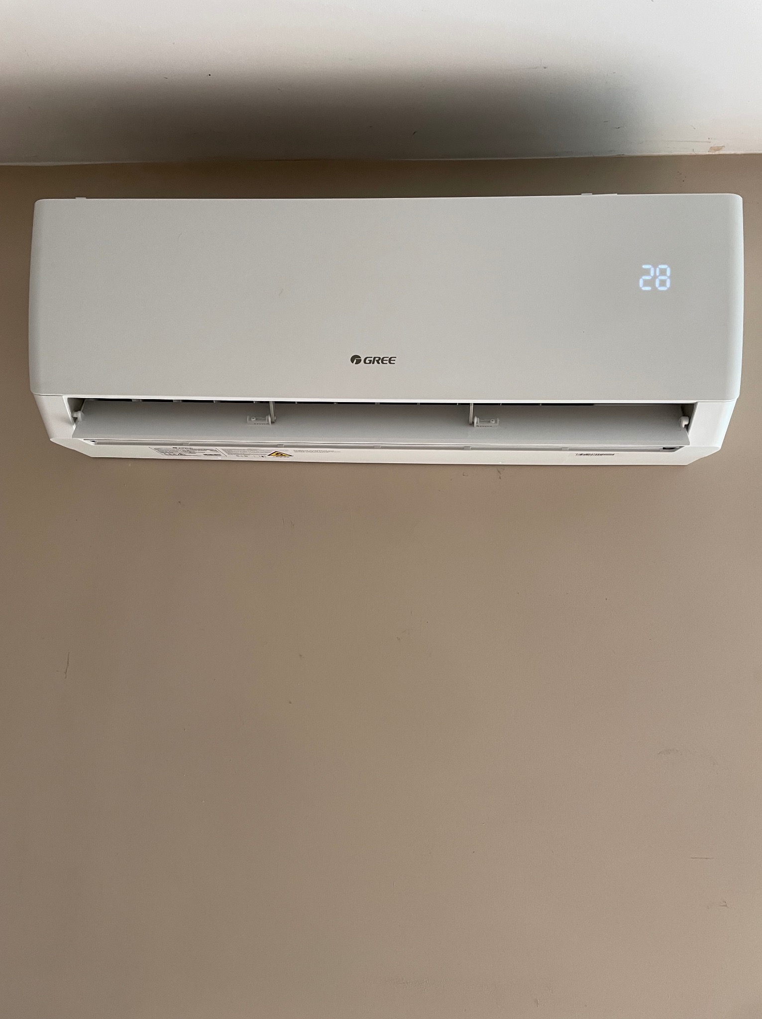 Biała klimatyzacja marki Gree zamontowana na beżowej ścianie, wyświetlająca temperaturę 28 stopni.