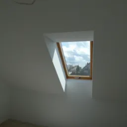 Poddasze z oknem dachowym, świeżo wyszpachlowane i pomalowane na biało, widok na osiedle domów jednorodzinnych za oknem.