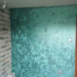 Ściana z dekoracyjną, turkusową farbą strukturalną, imitującą efekt aksamitu, z fragmentem ściany wykończonej kamieniem dekoracyjnym po lewej stronie.