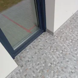 Ułożona mozaika z płytek w odcieniach szarości i beżu na balkonie, widok z góry, z fragmentem szarej ramy okiennej i białej ściany. Drobne elementy tworzą wzór.