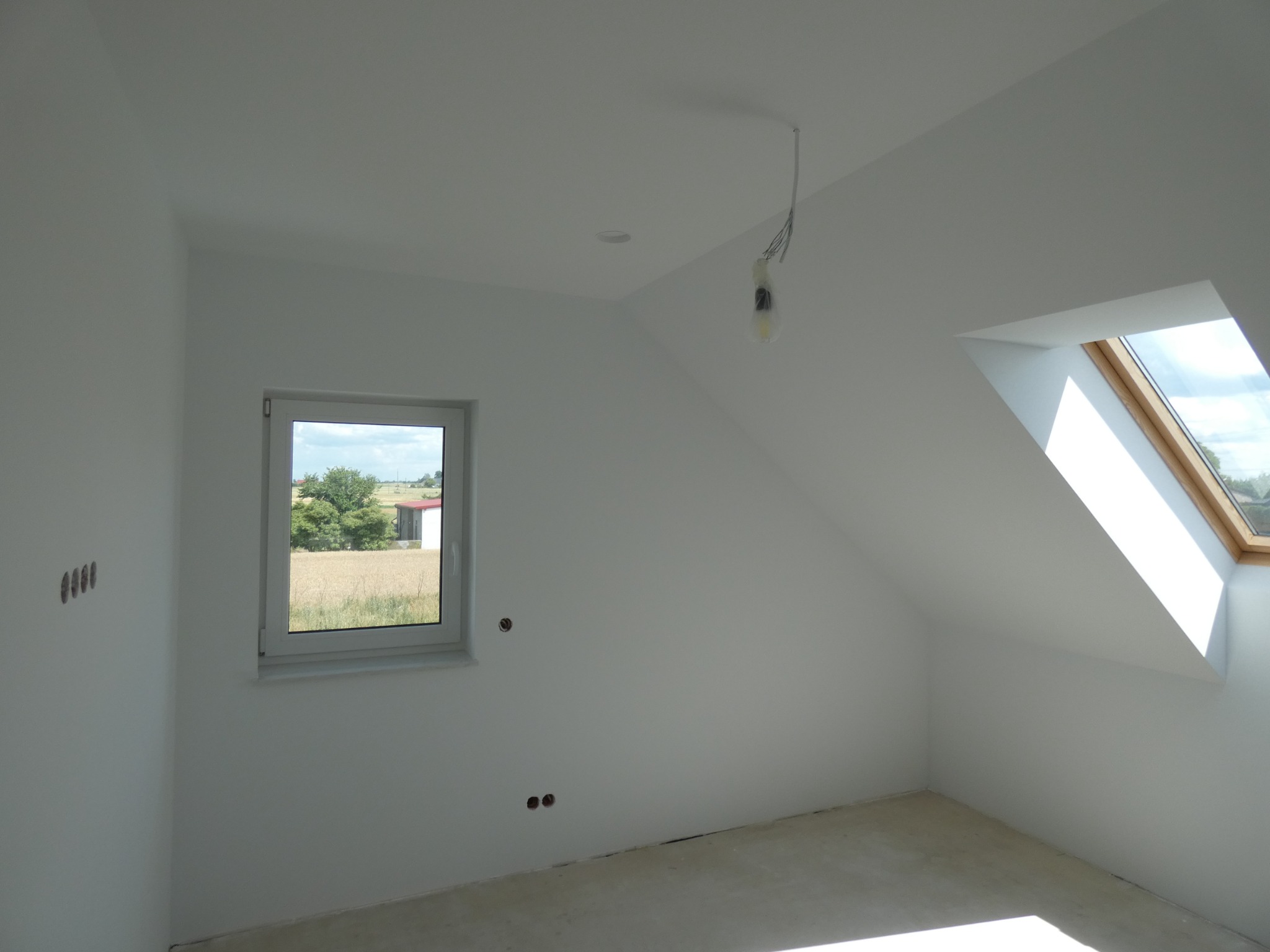 Poddasze w stanie deweloperskim z oknem dachowym i widokiem na pole, surowe ściany przygotowane pod malowanie, wystające kable elektryczne.