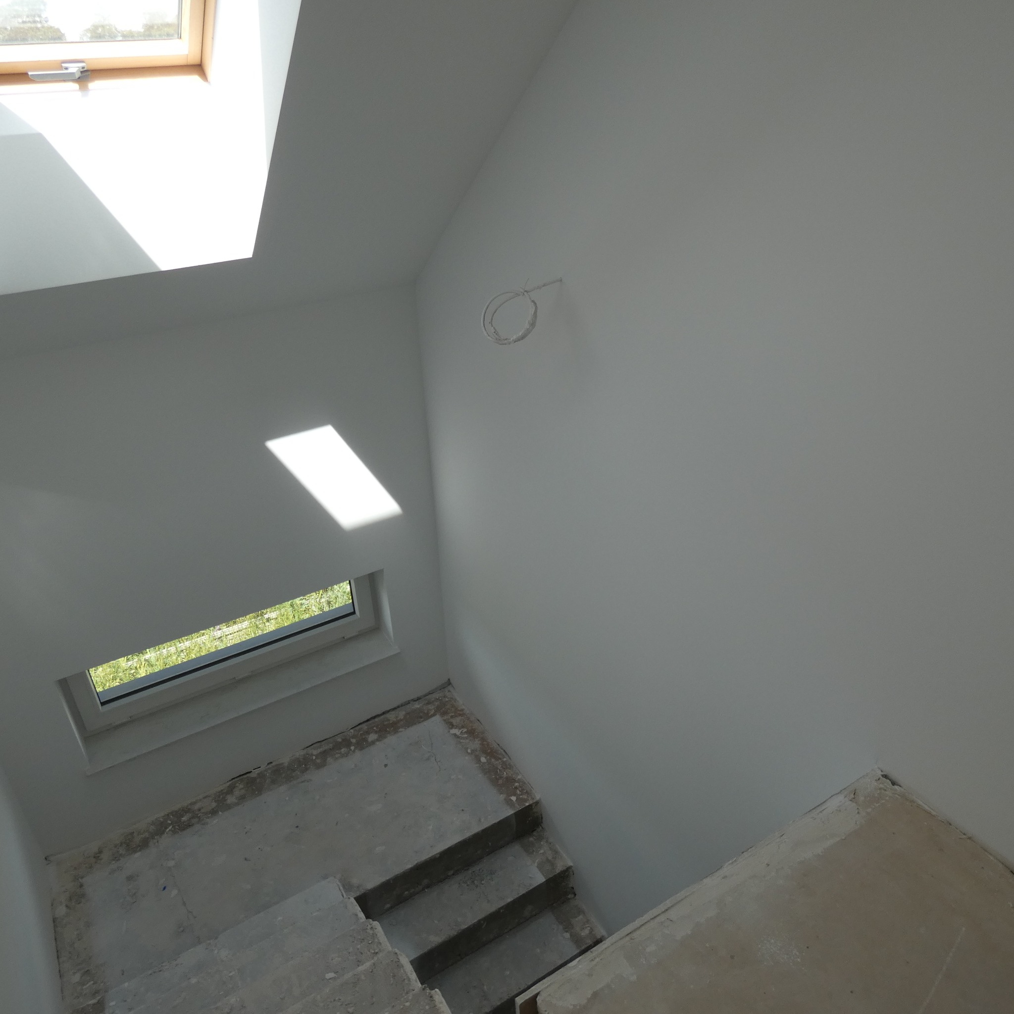 Surowe schody betonowe prowadzą w dół, ściany pokryte świeżo nałożoną gładzią, widoczne kable elektryczne wystające ze ściany, okno dachowe wpuszczające światło.