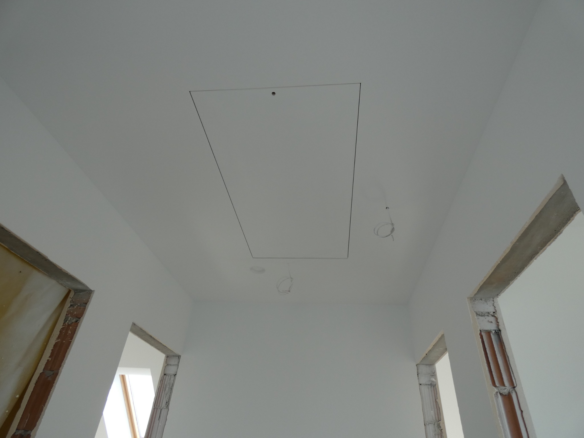 Białe wnętrze w trakcie prac wykończeniowych: widok na sufit z klapą rewizyjną i niedokończone ościeżnice drzwiowe z widoczną cegłą.