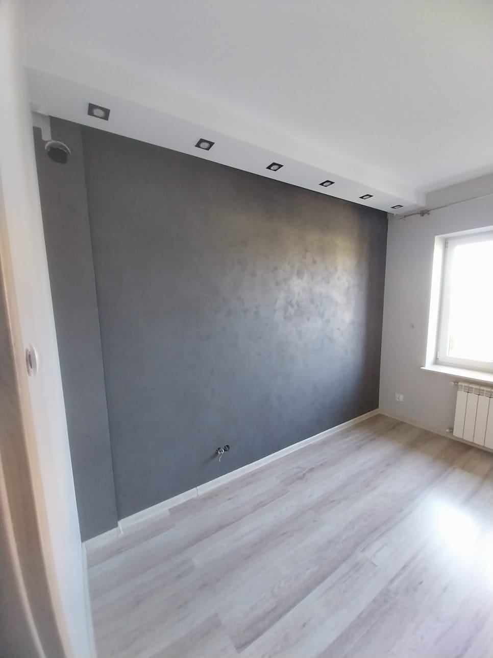 Pokój po remoncie: ściana w odcieniach szarości z dekoracyjną fakturą, jasna podłoga panelowa, białe listwy przypodłogowe, sufit z oświetleniem punktowym, okno z grzejnikiem.